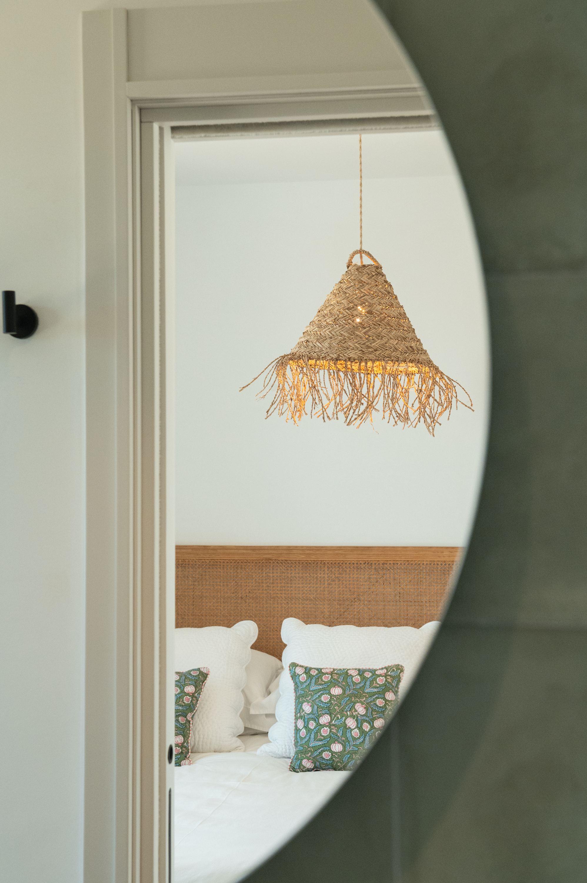 Reflection of a bedroom seen through a mirror, featuring a bed with white pillows and floral-patterned throw pillows, a woven straw pendant light, and a wooden headboard.