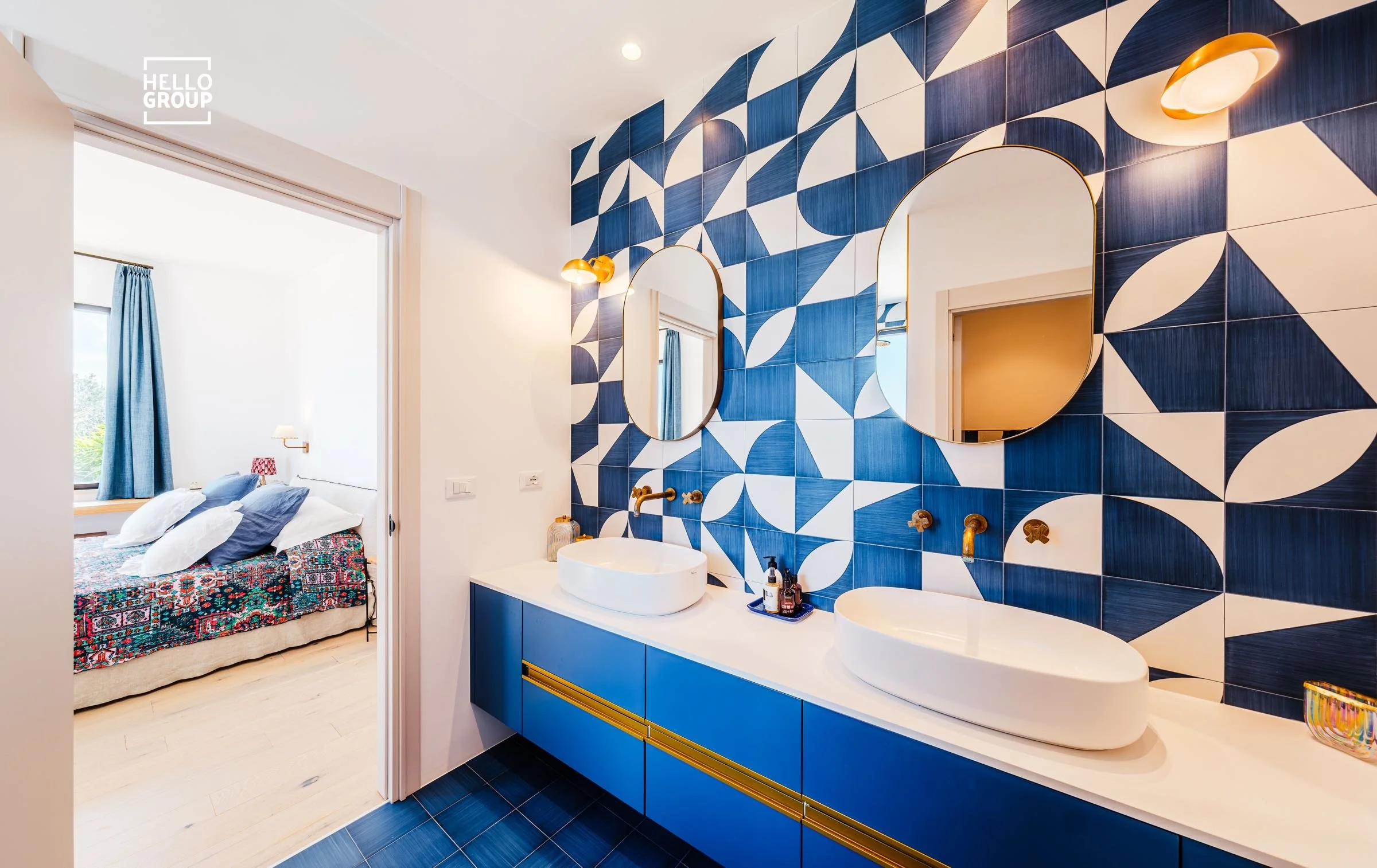 Modern bathroom with two white vessel sinks on a white vanity, gold fixtures, an abstract blue and white tile wall, two oval mirrors, and warm lighting.