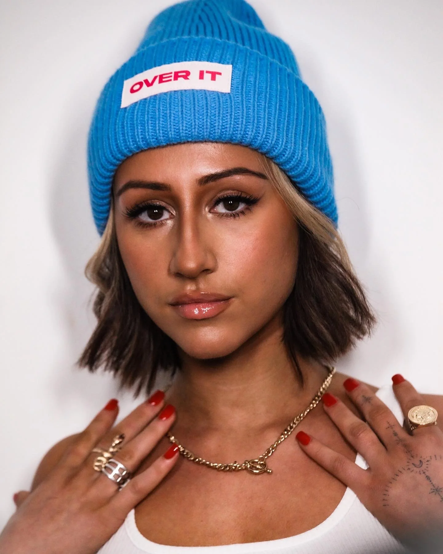 A woman with shoulder-length brown hair, wearing a blue knit beanie with a red and white 'OVER IT' patch, a gold chain necklace, rings, and red nail polish, posing with her hands on her shoulders against a white background.