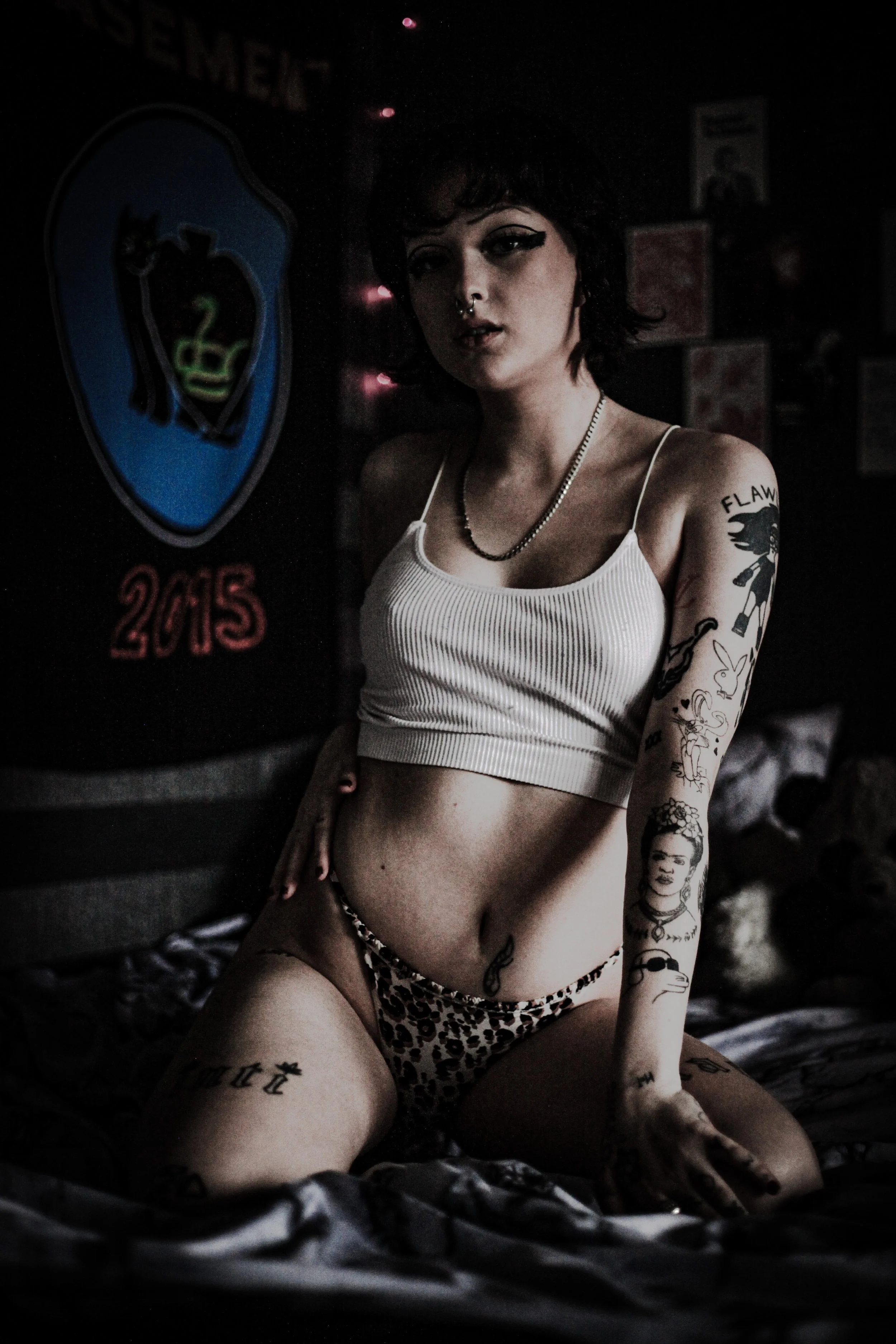 A young woman with tattoos, short dark hair, and piercings poses in a dimly lit room wearing a white crop top and leopard print underwear, with a neon sign reading "2015" on the wall behind her.