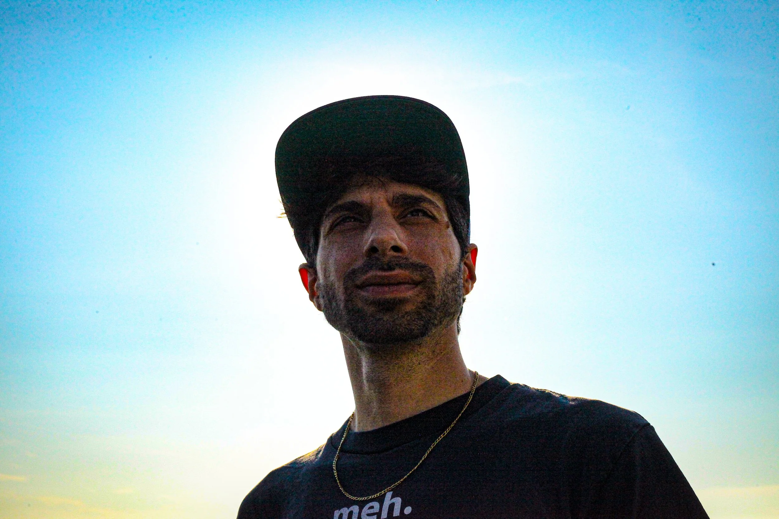 Young man wearing a black cap and T-shirt, standing outdoors with the sun behind him, looking into the distance.