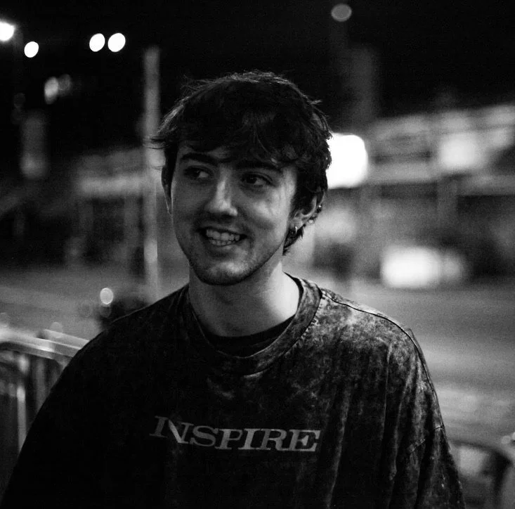 A young man with dark hair and earrings smiling at night, wearing a T-shirt that says 'INSPIRE', in an outdoor setting with blurred lights in the background.