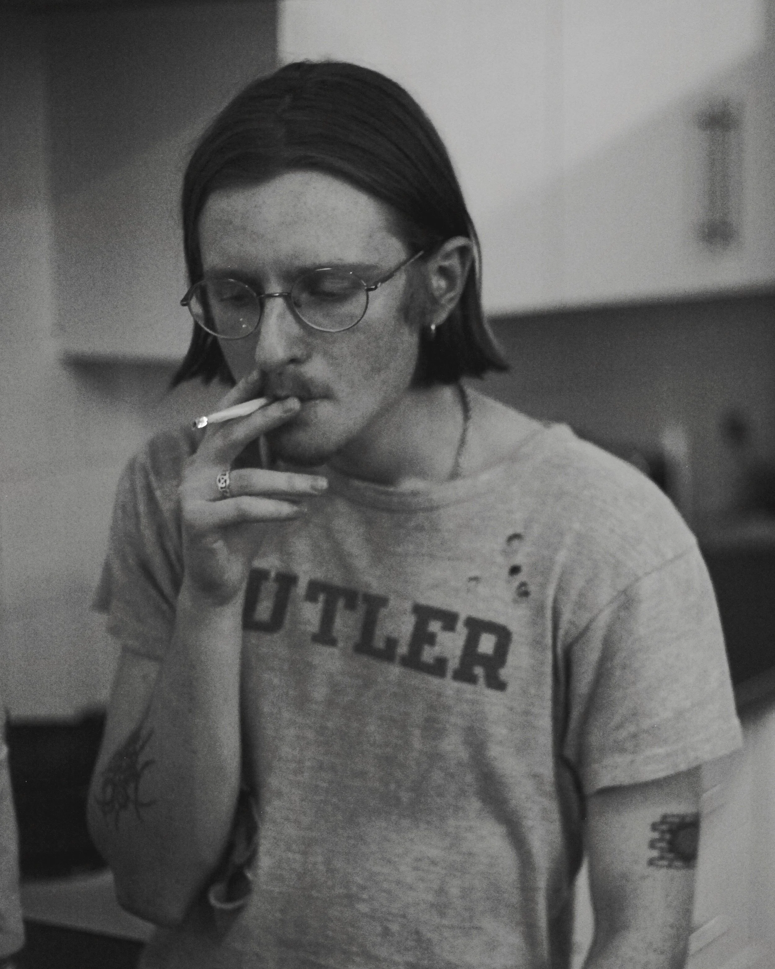 Black and white photo of a young person with long hair, glasses, and tattoos, smoking a cigarette.