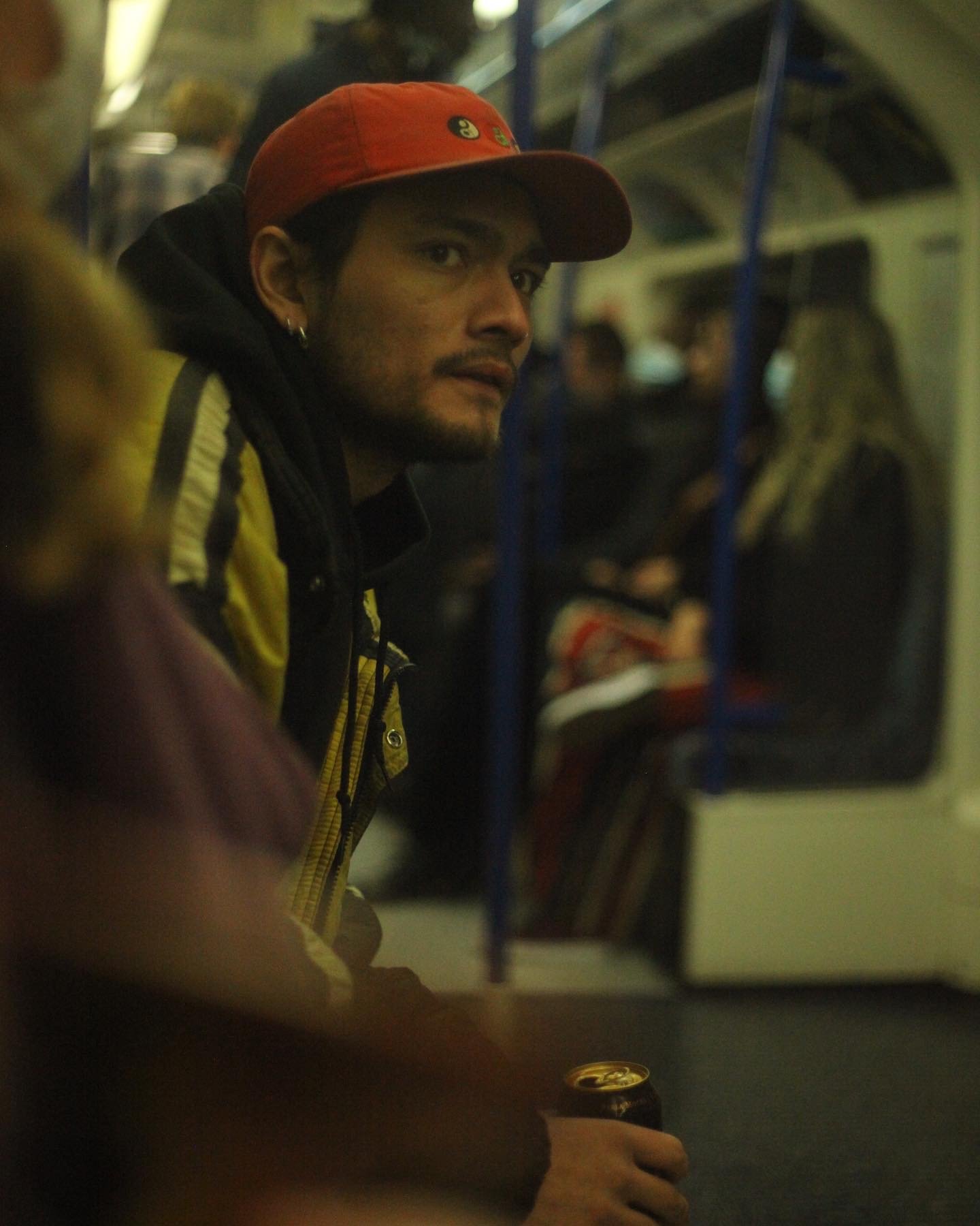 A man with a red baseball cap, earrings, and a yellow jacket sitting on a subway train holding a can of soda.