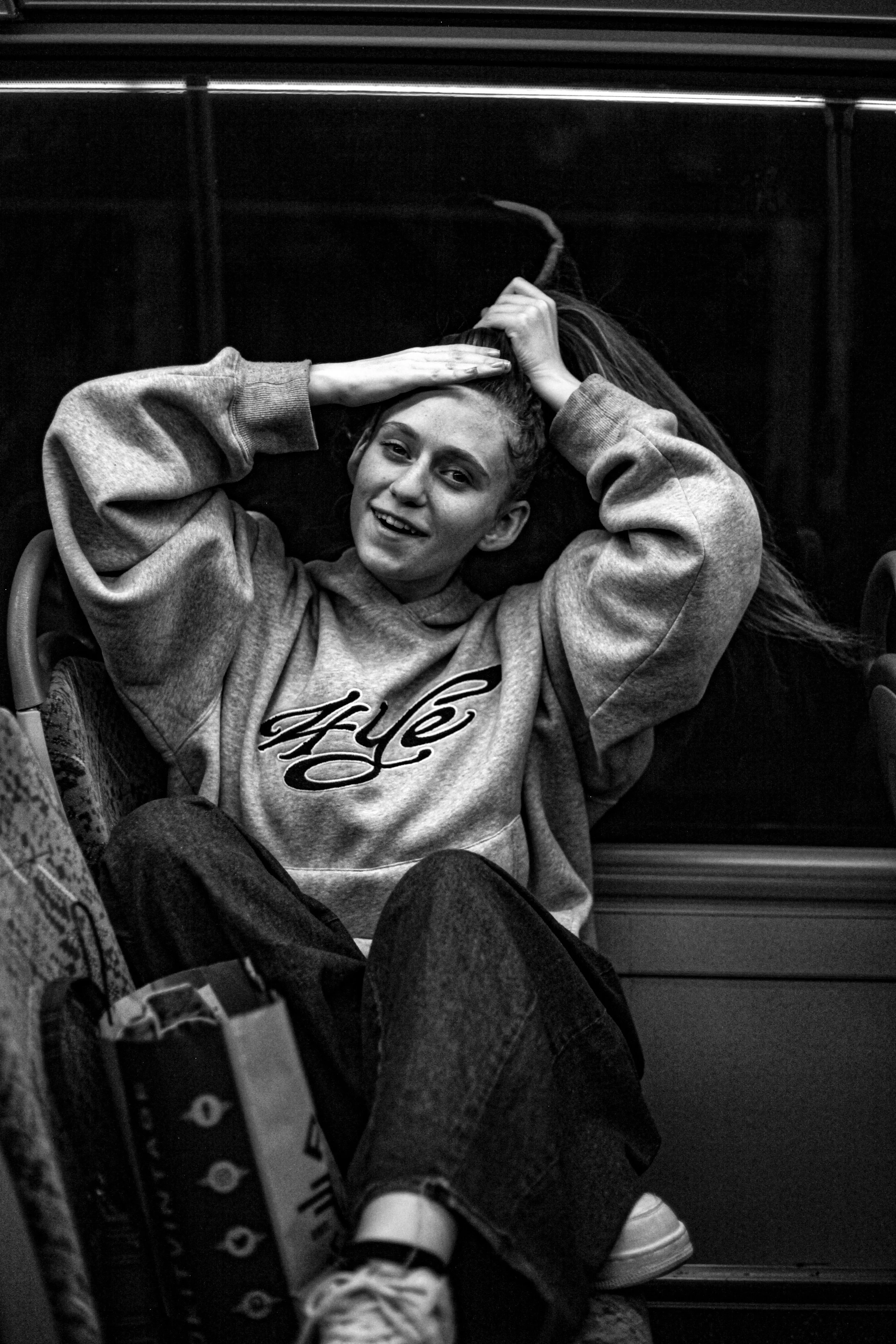 A young woman in a hoodie  smiling, sitting on a bus with her hand holding her ponytail, and other passengers and seats visible around her.