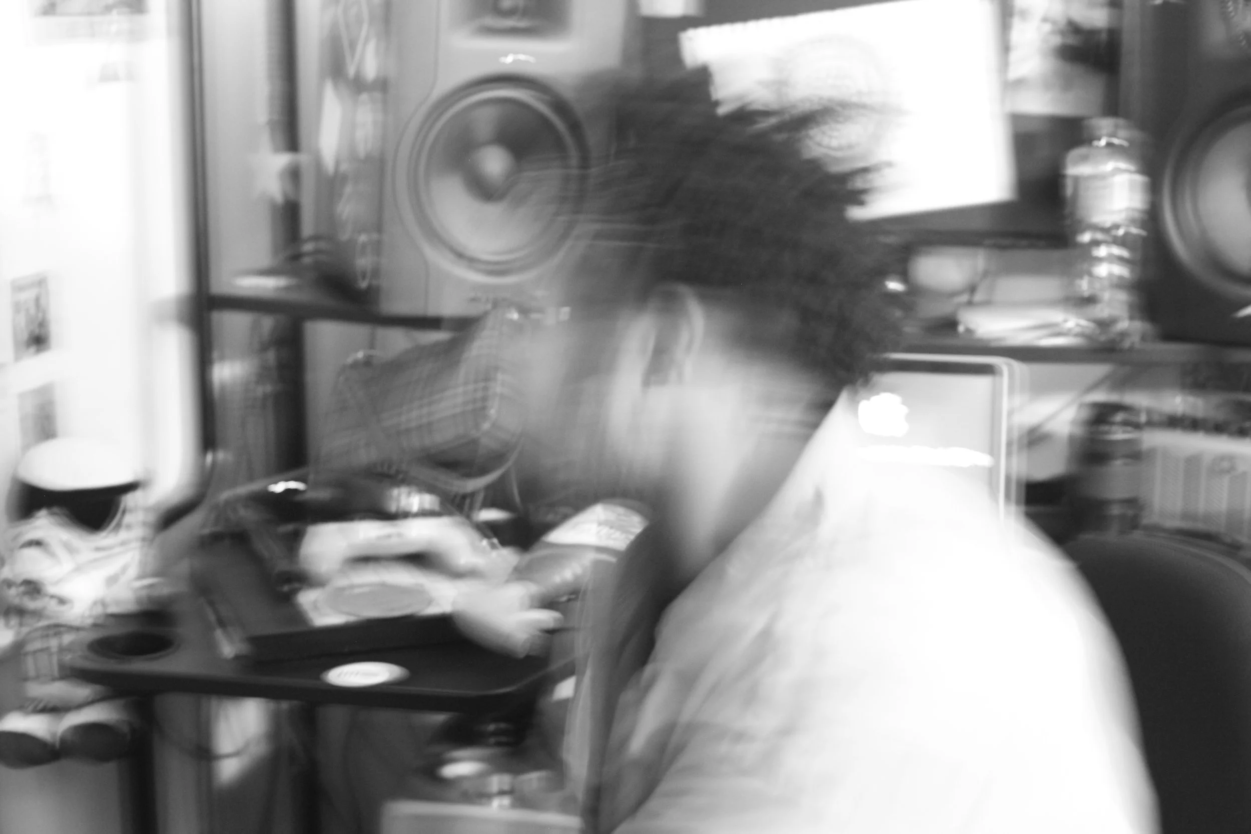 Blurred black and white photo of a person in plaid shirt working at a desk with speakers, a laptop, and other objects.