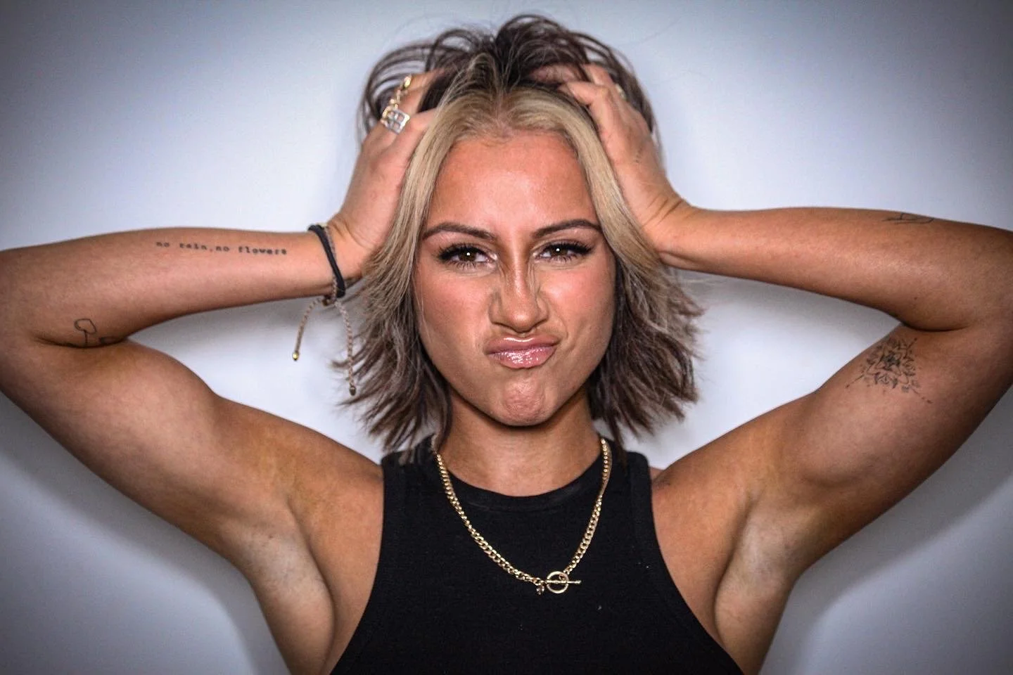 A woman with short, wavy blonde hair and tattoos on her arms is holding her head with both hands and making a quizzical face.