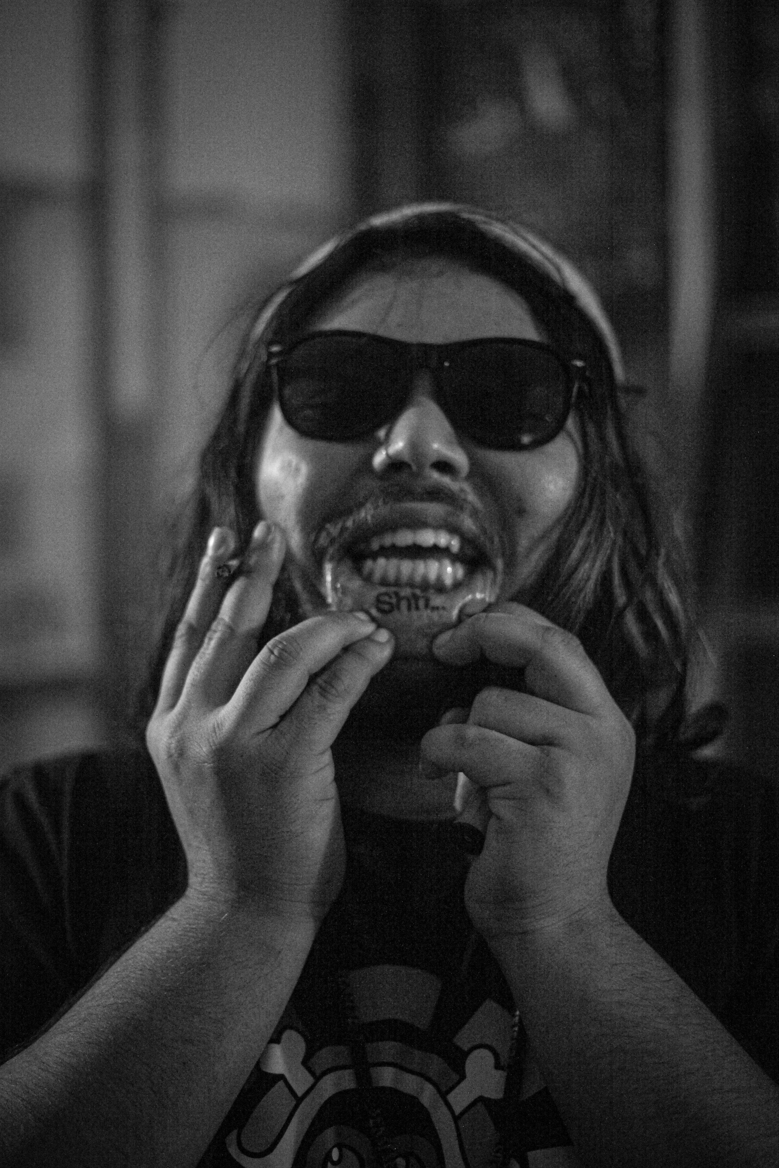 A person wearing sunglasses revealing a tattoo on the inside of his lip that says "shh..." in a black and white photo.