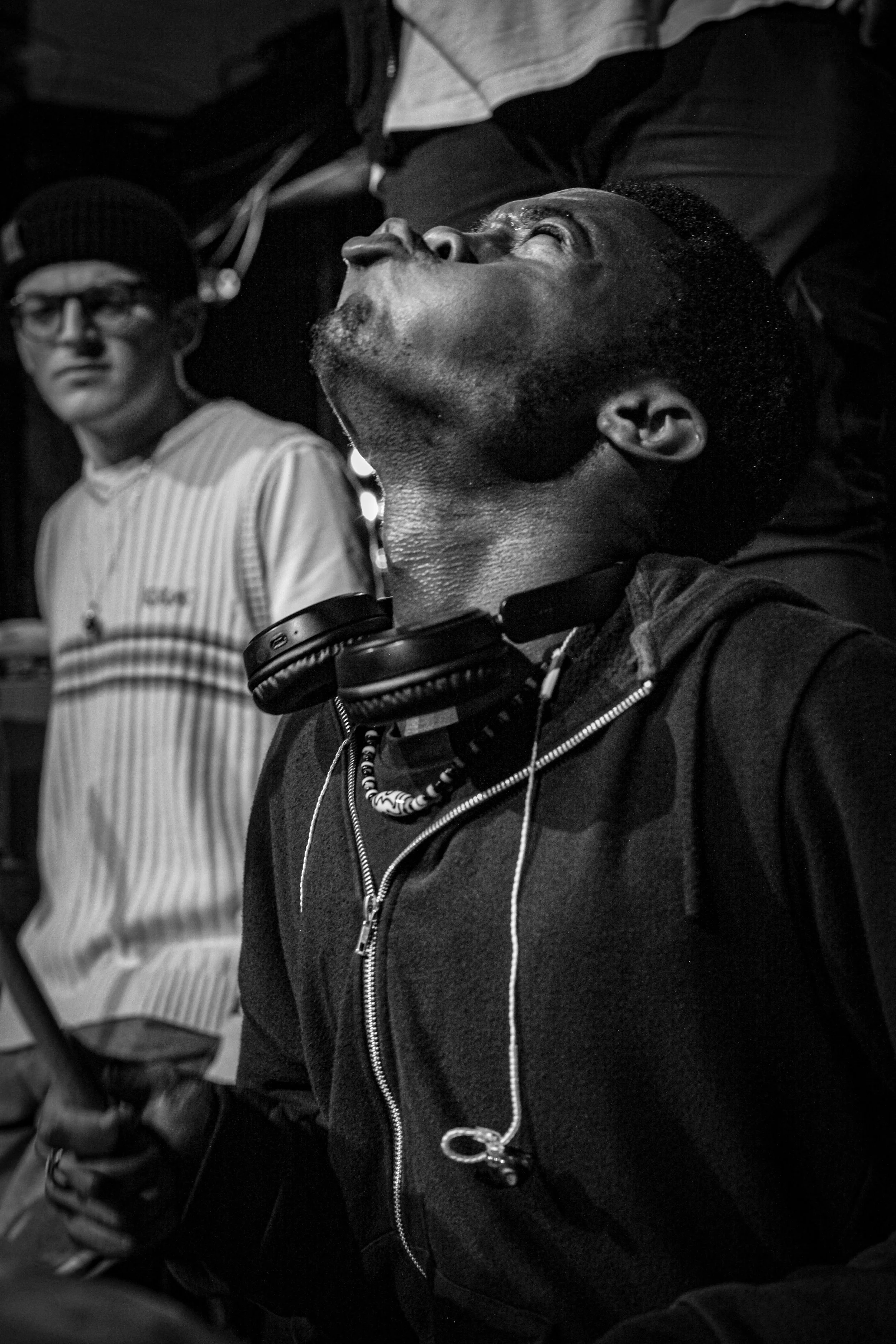 A black and white photo of a person with headphones around their neck, eyes closed, head tilted back. A second person in the background is standing, wearing glasses, a striped shirt, and a beanie.