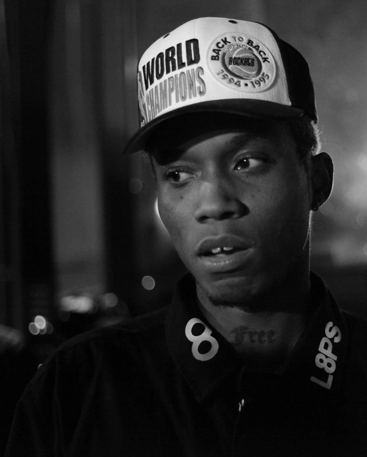 A young man wearing a baseball cap and jacket with a "back to back championship" logo, looking to the side, in black and white.