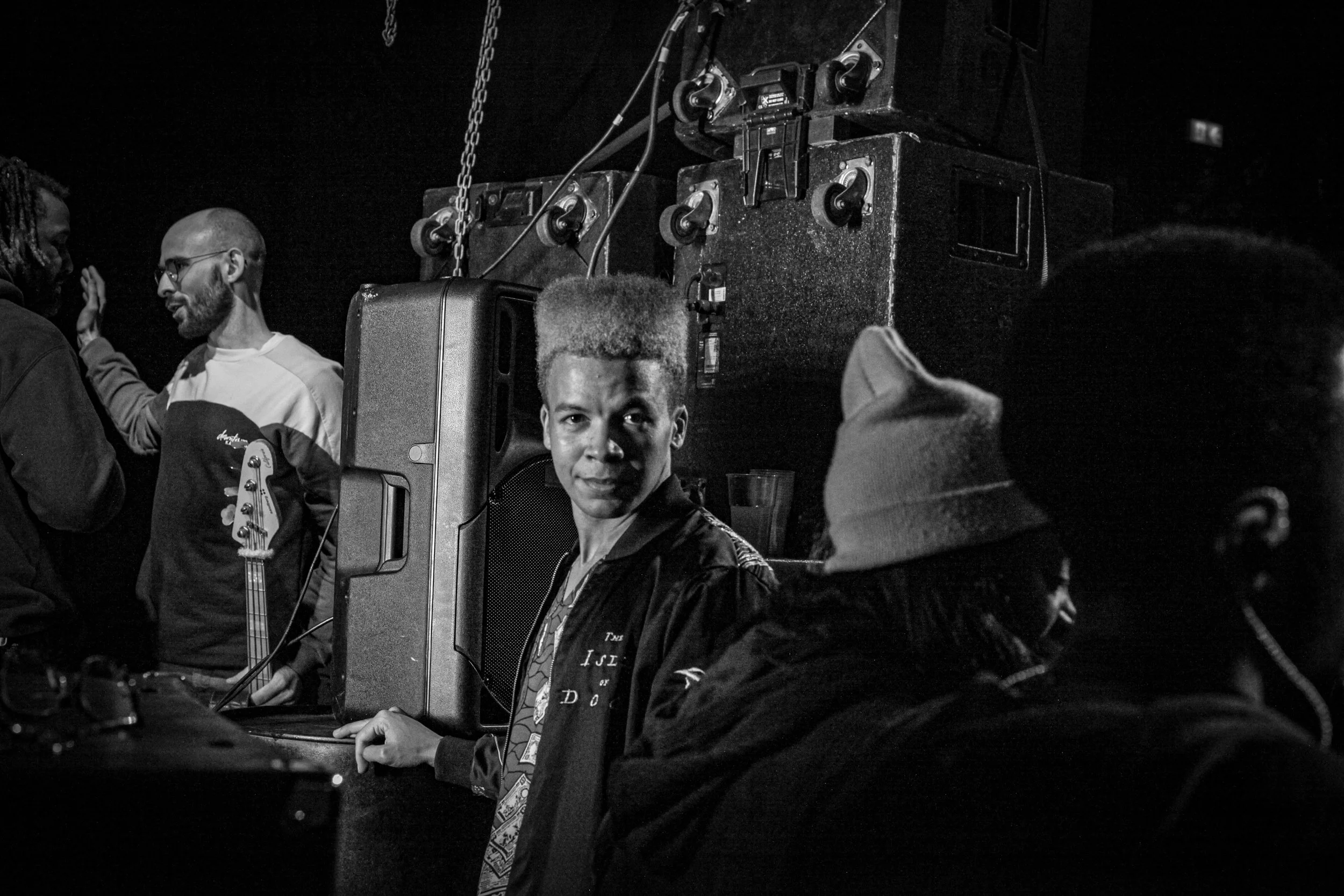 Black and white photo of people at a social gathering, with a woman in the center looking at the camera, surrounded by others engaged in conversation, and large speakers in the background.