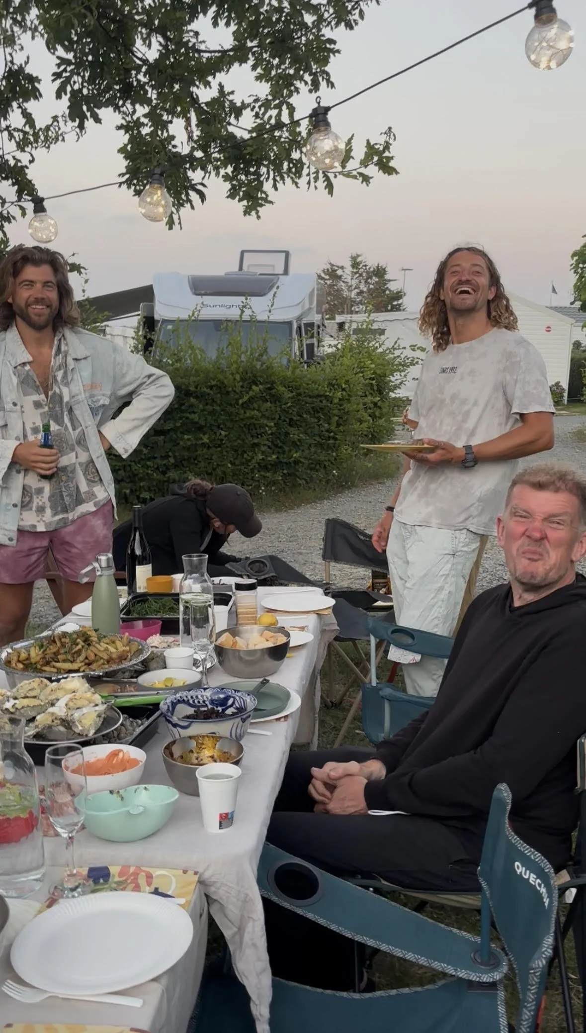 People celebrating around a table outdoors with food, drinks, and string lights hanging above during sunset.