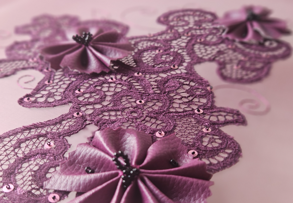 A close-up view of pink lace embroidery with satin ribbon flowers and small pink sequins on a pink background.