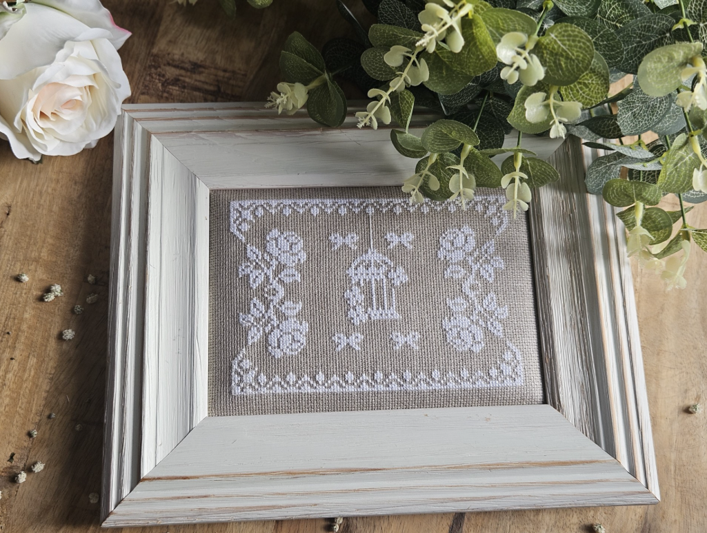 Framed cross-stitch embroidery with a birdcage, flowers, and bows, placed on a wooden surface with white flowers and green foliage.