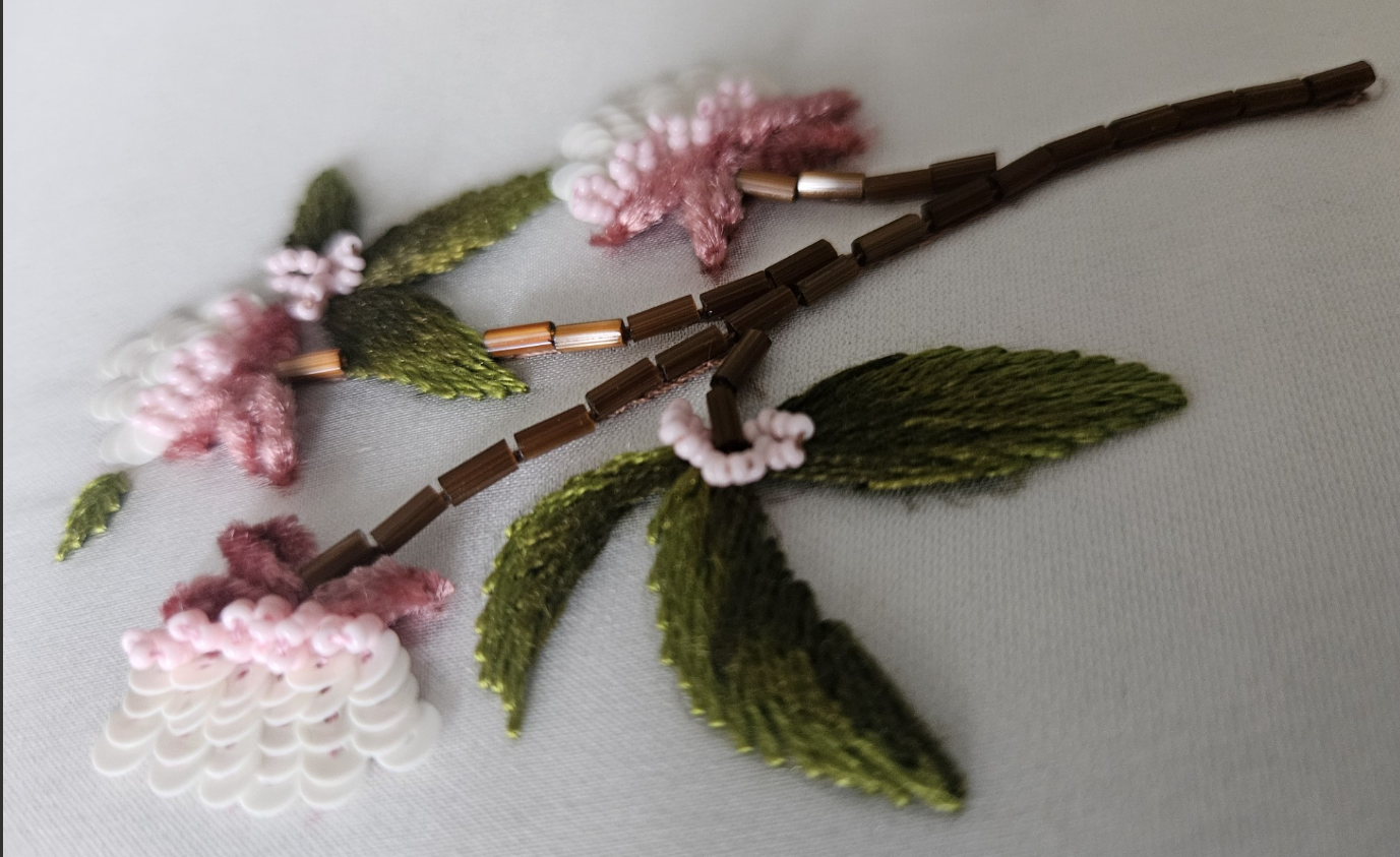Bead embroidery of a pink and white flower with green leaves on fabric.