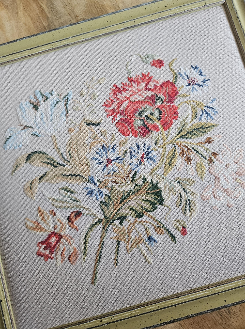 Cross-stitched floral bouquet with red, blue, beige, and white flowers in a frame on a wooden floor.