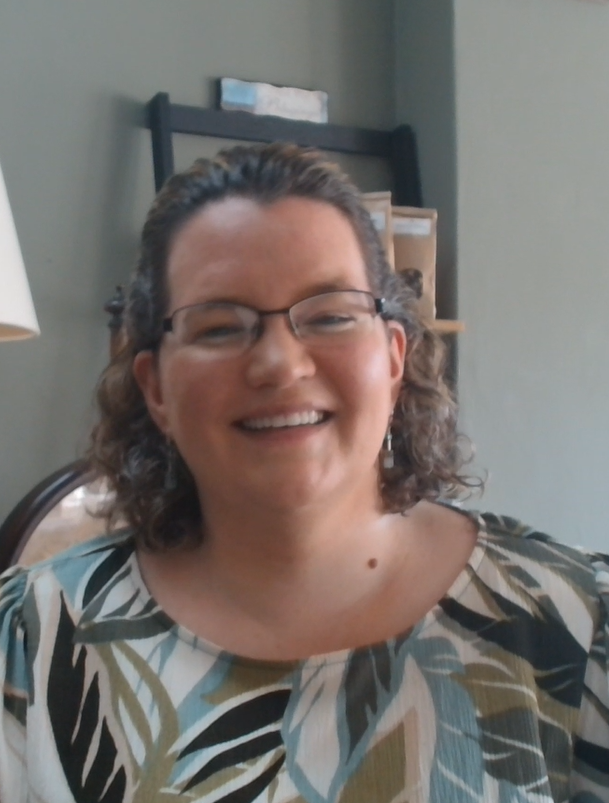 A woman smiling, wearing glasses and a patterned blouse, sitting indoors with a bookshelf in the background.