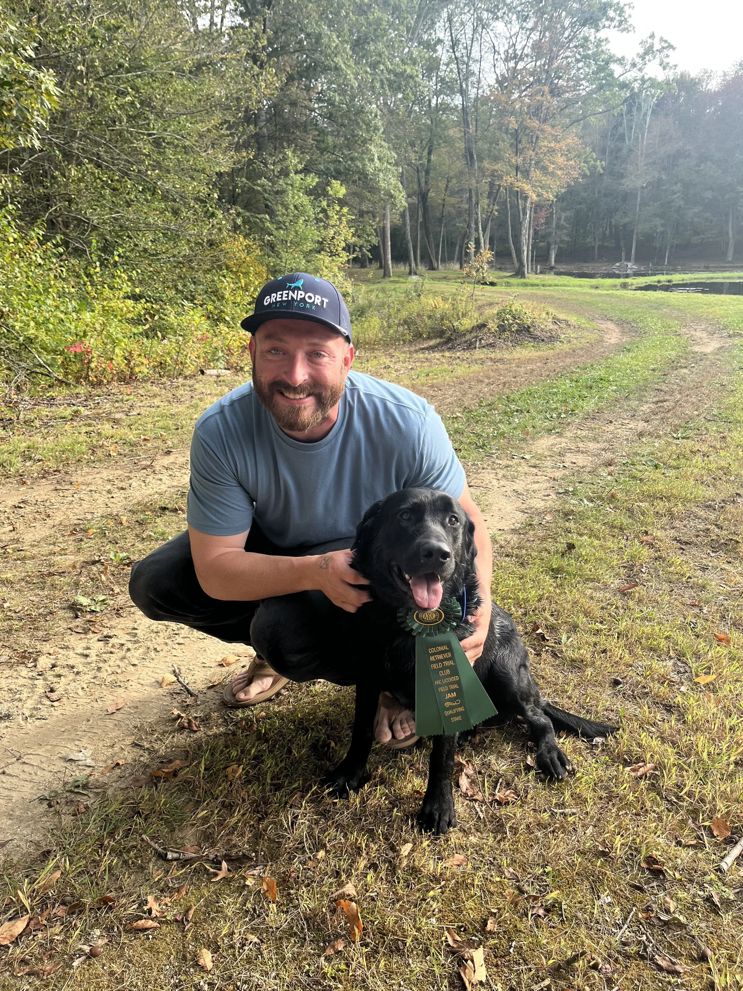 A man squatting outdoors with a black Labrador retriever, both smiling. The man is wearing a blue t-shirt and a black cap with an embroidered logo. The dog is wearing a green ribbon with gold text and a badge, indicating it is a trained field trial dog.