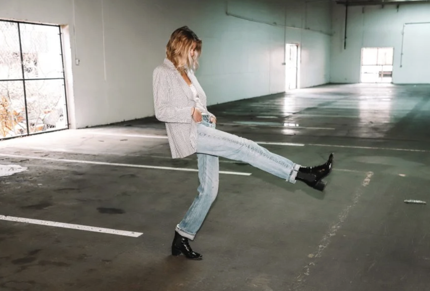 A woman standing in an empty parking garage with her left leg extended forward, wearing a checkered blazer, ripped jeans, and black boots, with her hands in her pockets and head slightly bowed.