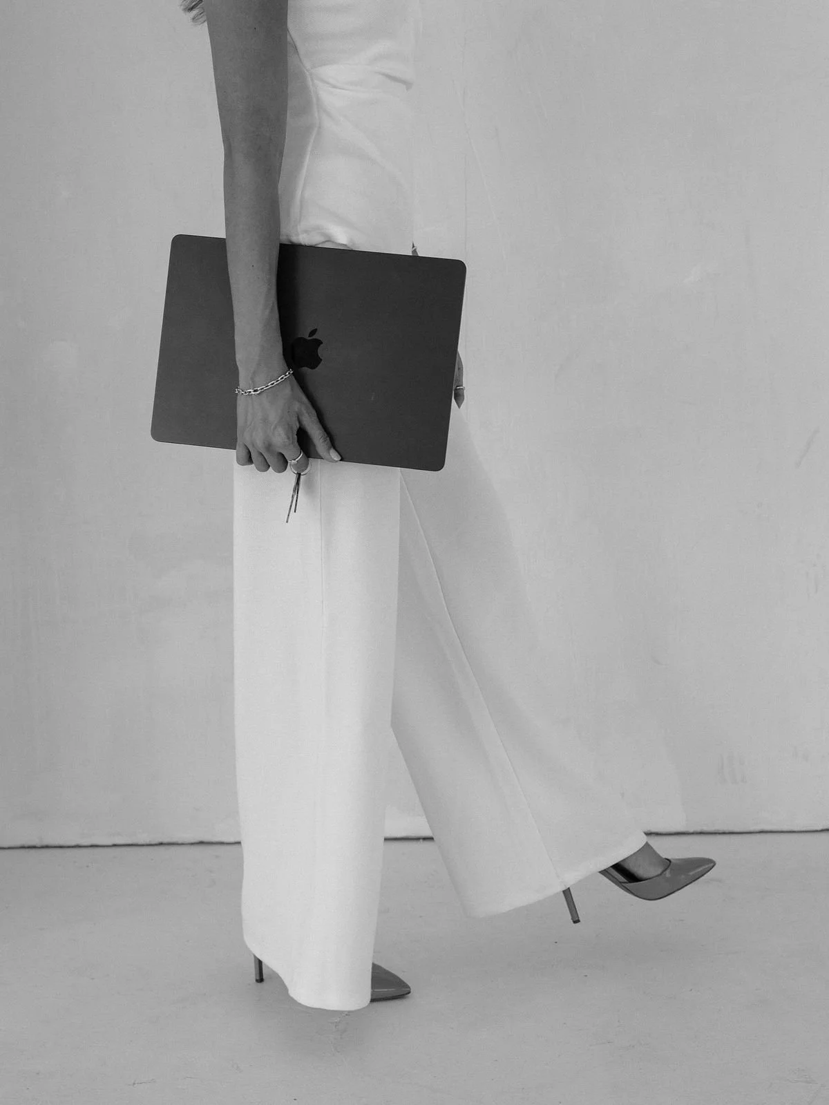 A person holding a closed MacBook laptop in their right hand while wearing white wide-leg pants and pointed high heels, standing against a plain wall.