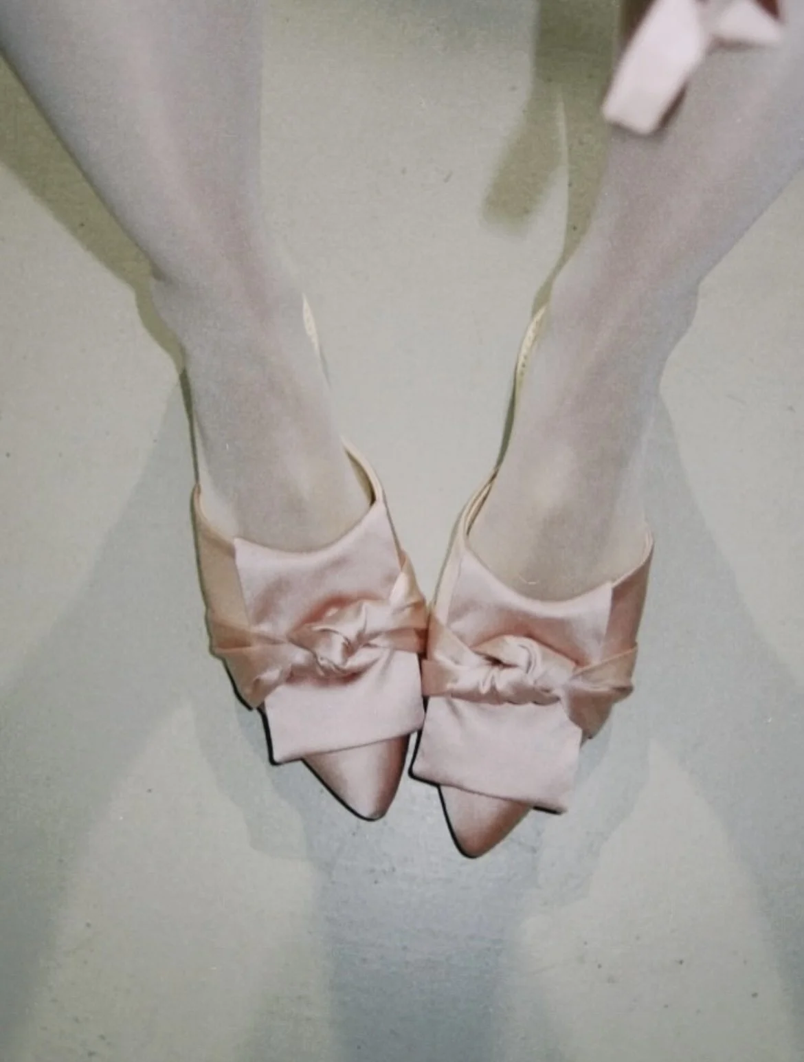 Pair of pink satin high heels with large bow decorations, photographed from above on a neutral gray surface.