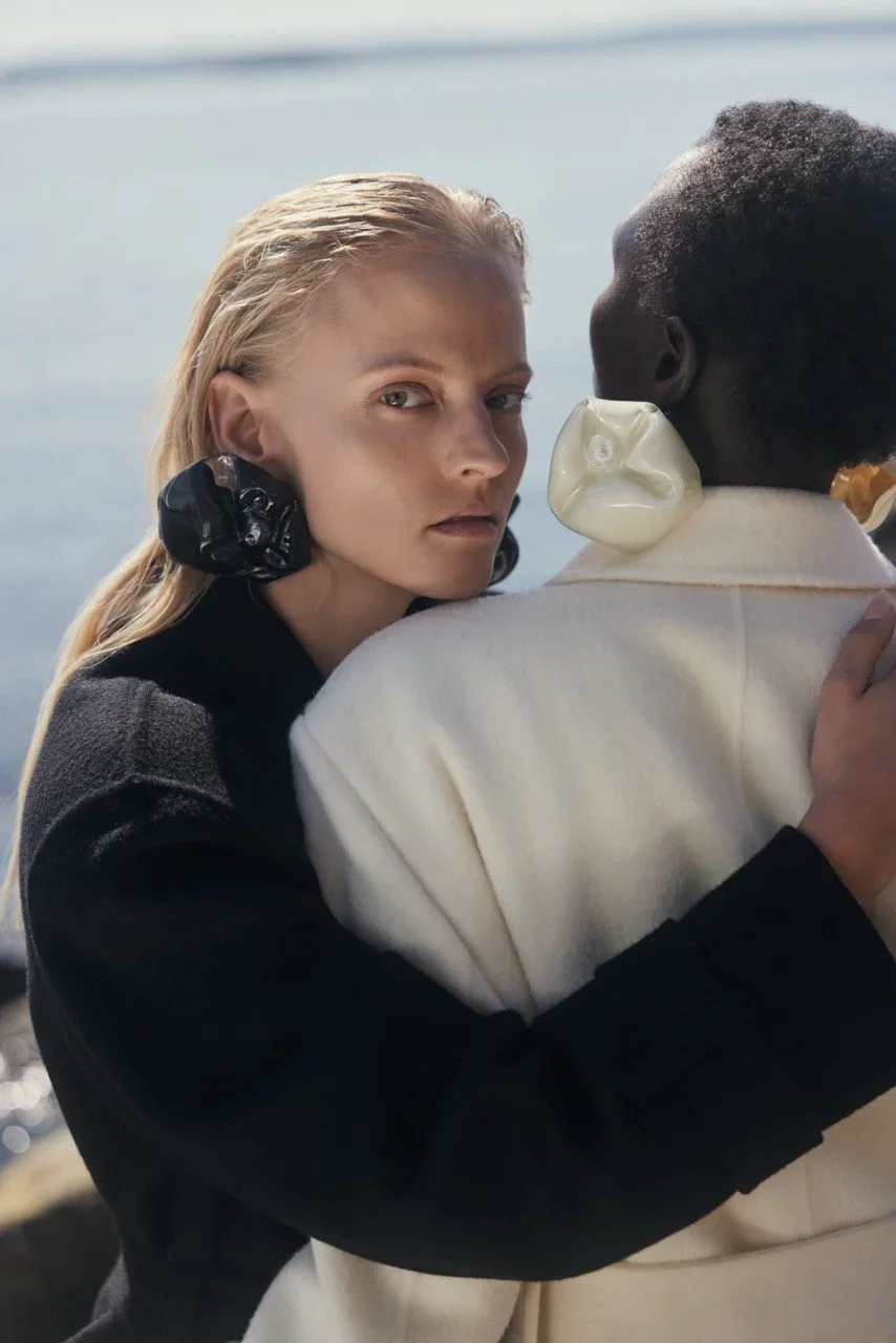 A woman with platinum blonde hair, wearing large black earrings, embraces a person with dark skin and short hair, who is wearing a white shirt. The person has a transparent ear device. They are outdoors near water.