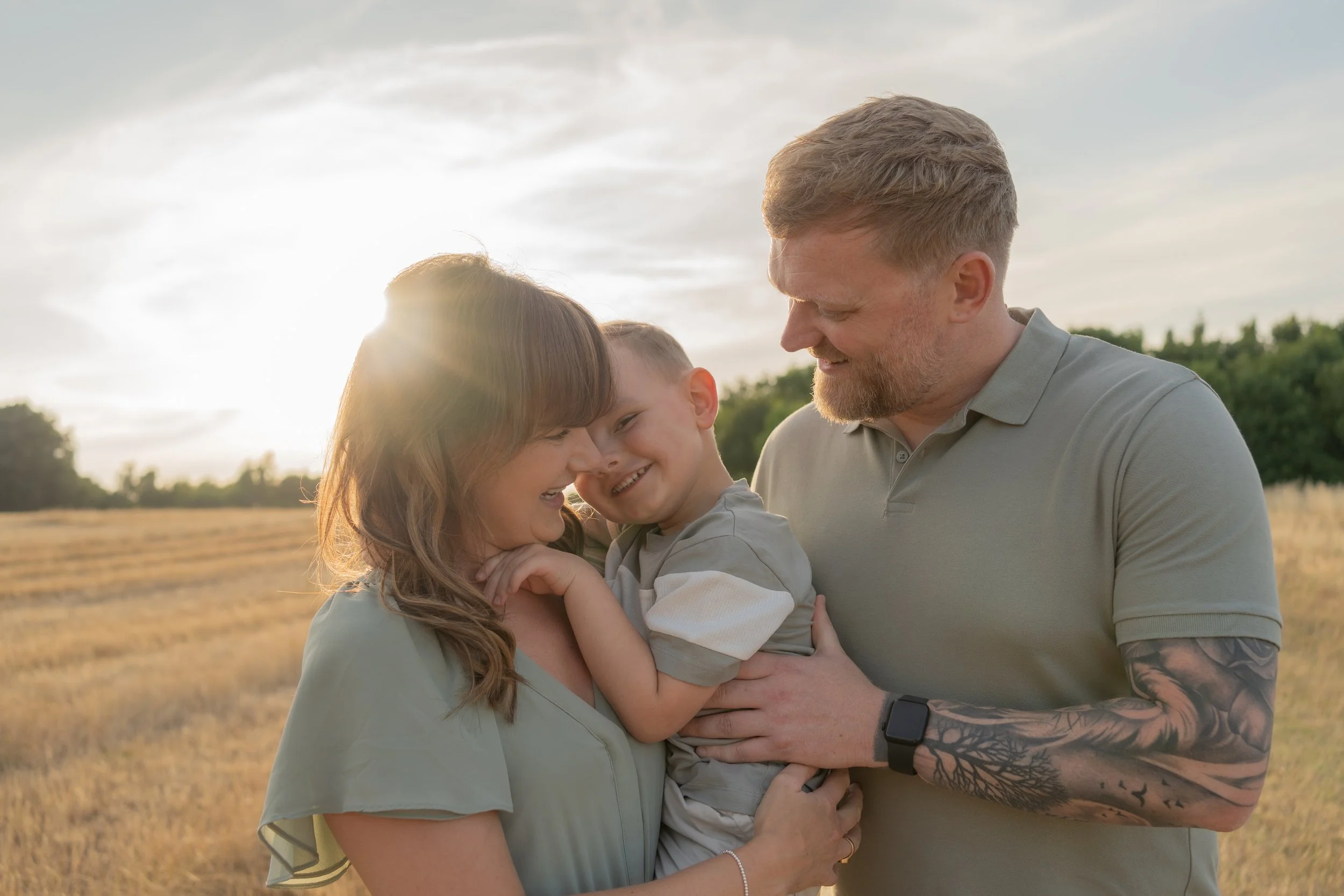 Family of three, a woman, a man, and a young boy, smiling and enjoying each other's company outdoors during sunset in a field.