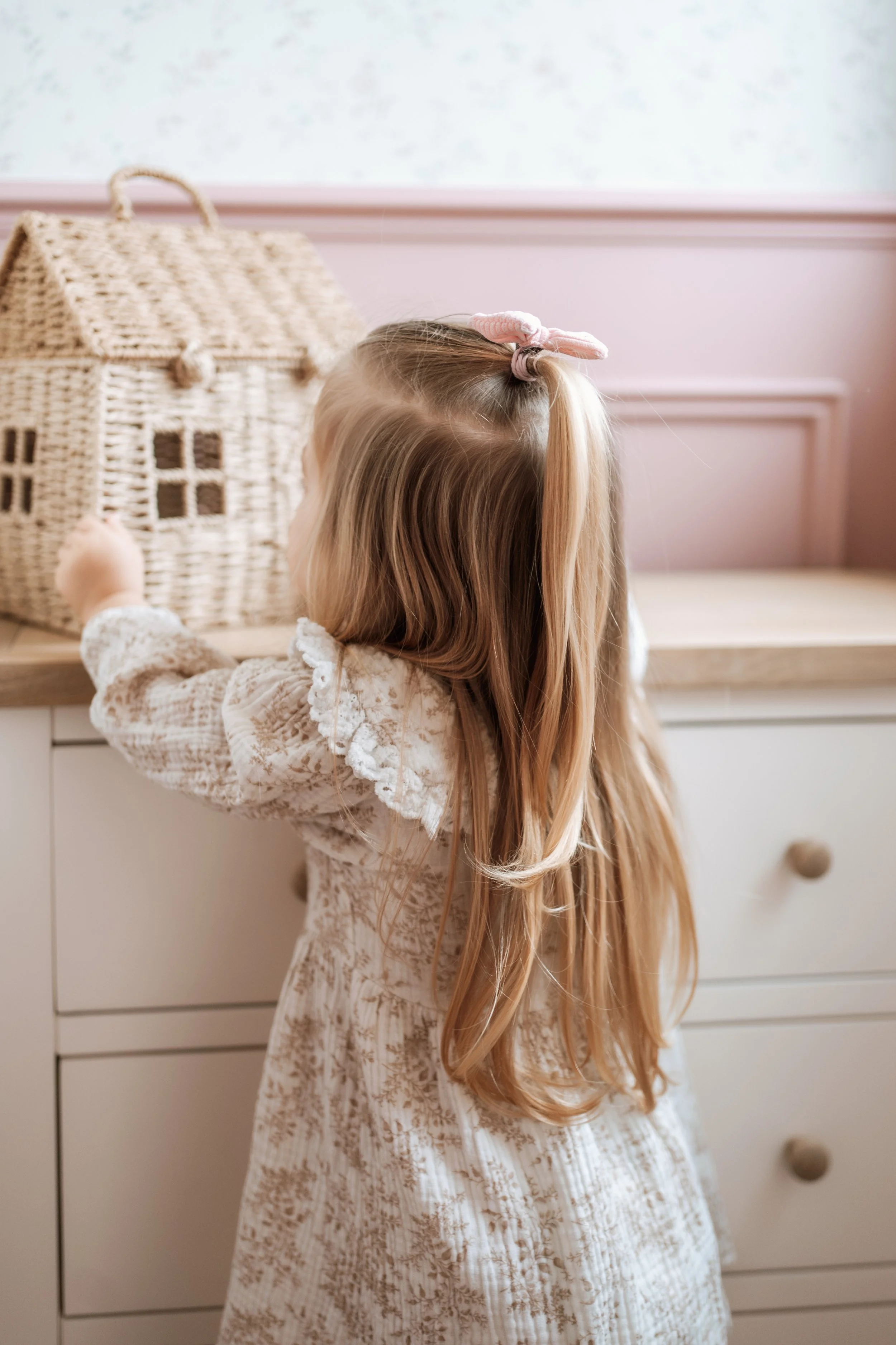 A young girl with long blonde hair tied with a pink bow, wearing a lace dress, standing at a white dresser with drawers, reaching for a woven basket on top.