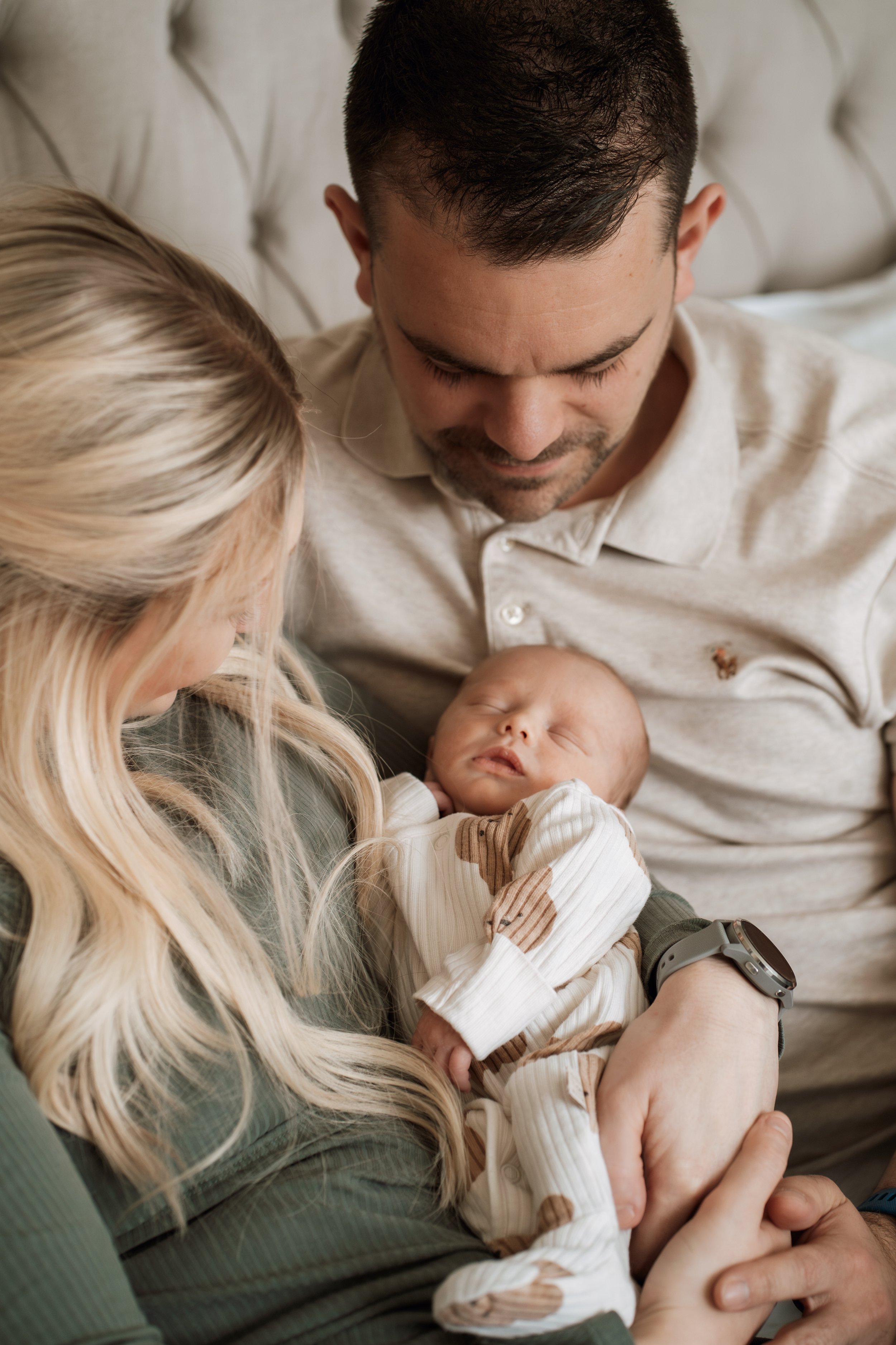 A man and a woman holding their sleeping baby, sitting on a bed.