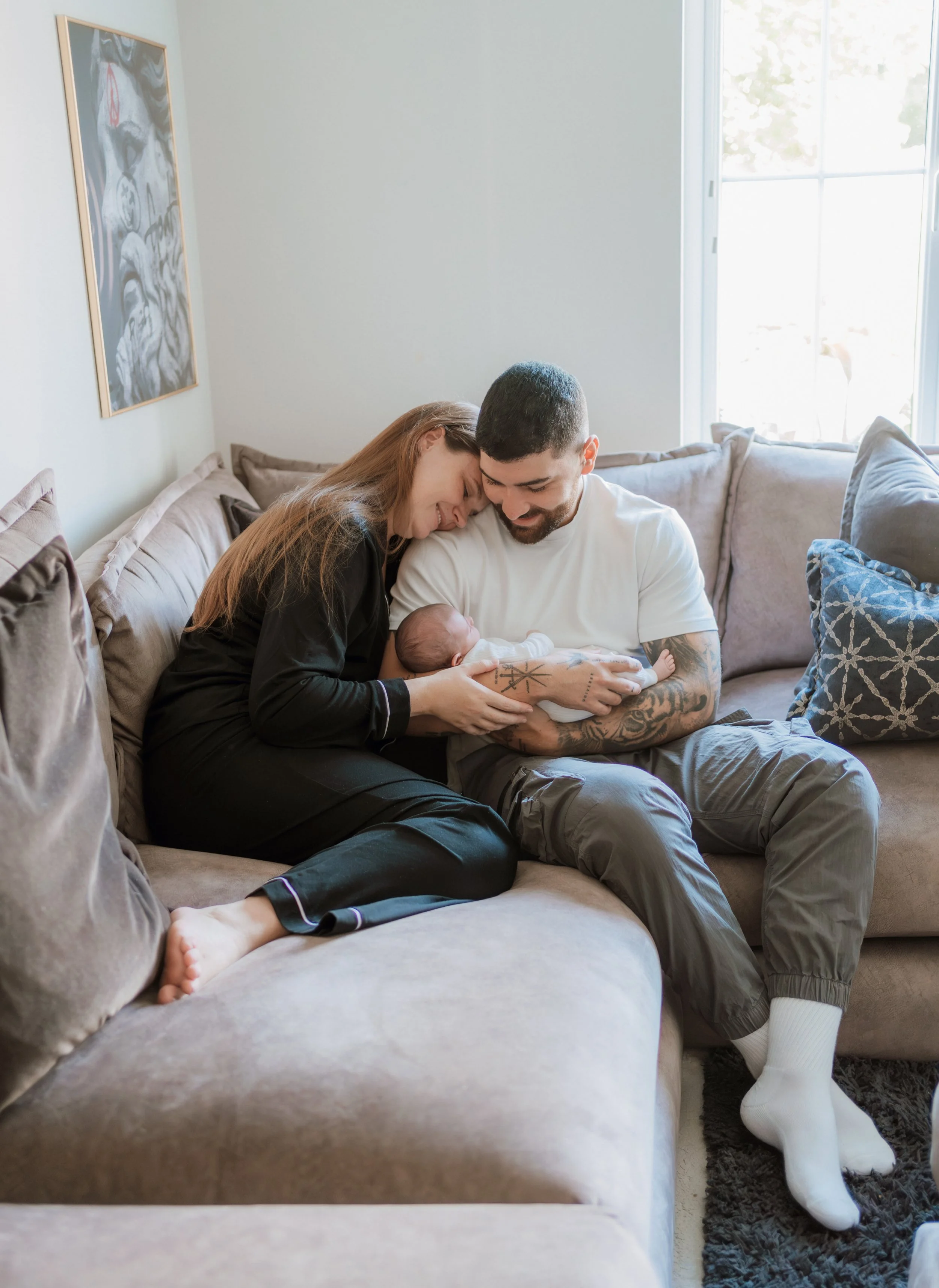 A family sitting on a couch in a living room, with a woman and man holding a newborn baby, smiling, and looking at the baby.
