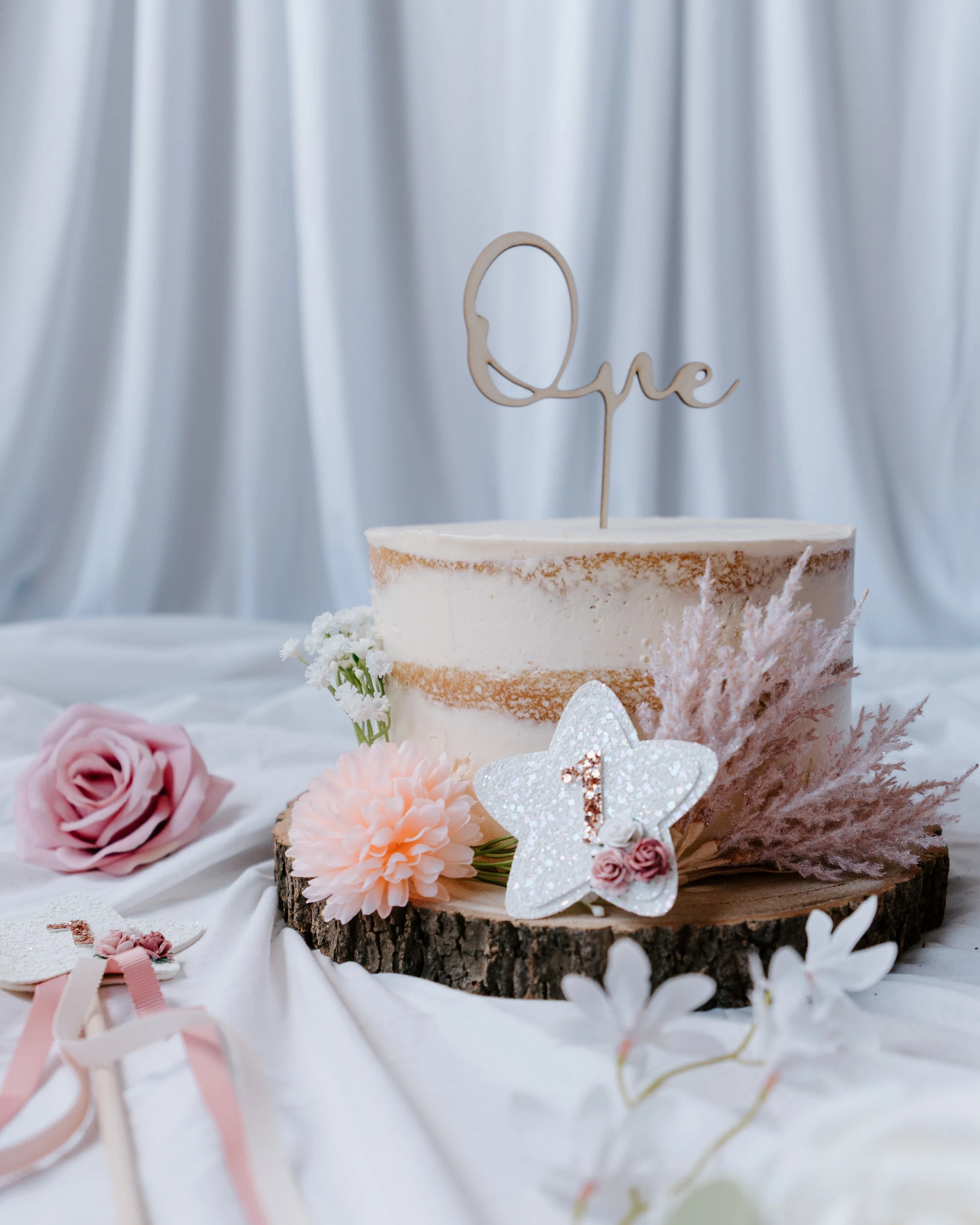A decorated birthday cake with a topper that says 'One,' surrounded by pink flowers, a white star-shaped decoration with a number one, and other floral and decorative accents on a wooden slice.