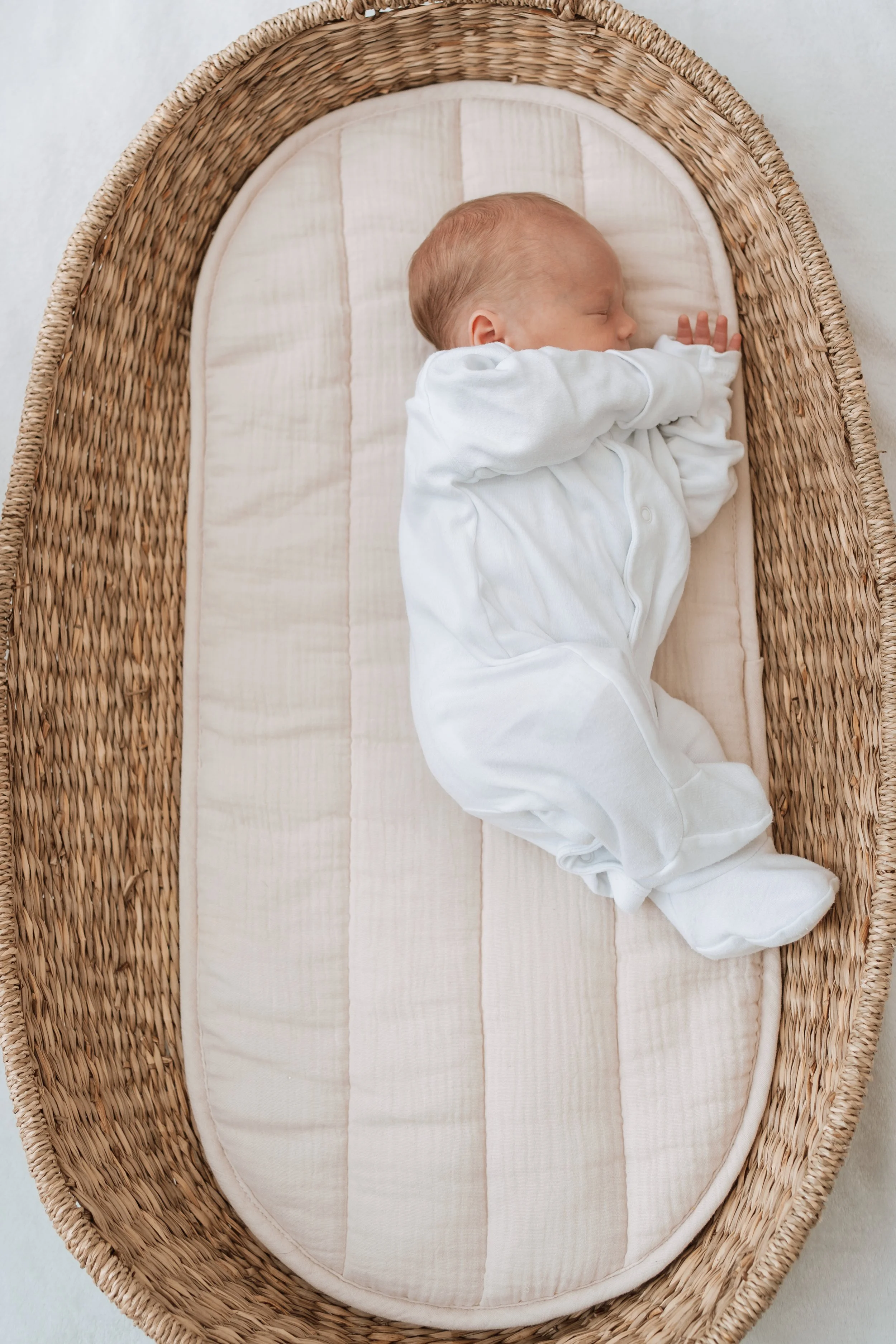 A sleeping baby in a white onesie lying on a soft blanket in a wicker bassinet.