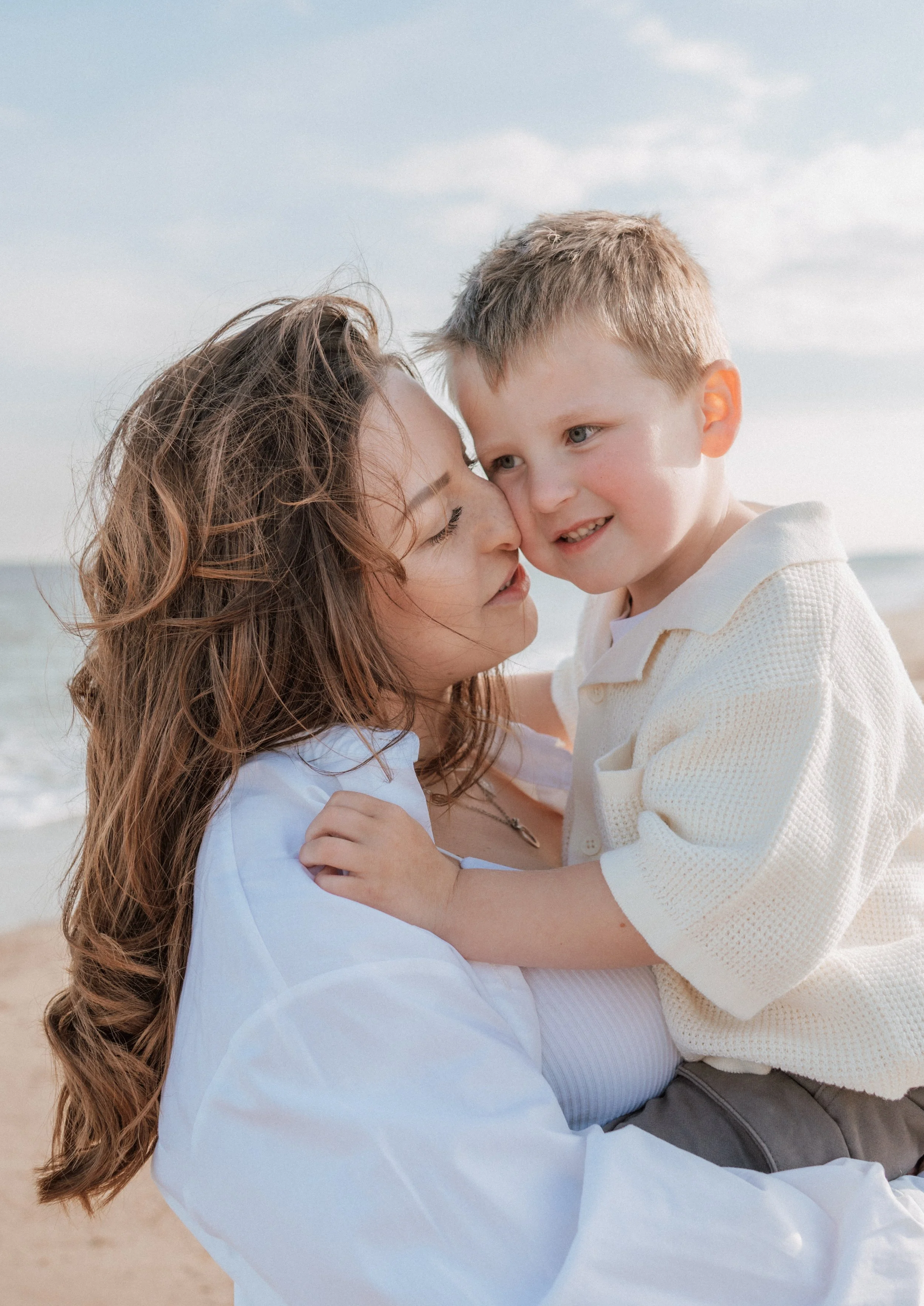 A woman holding a young boy at the beach, close to the shoreline with ocean waves behind them