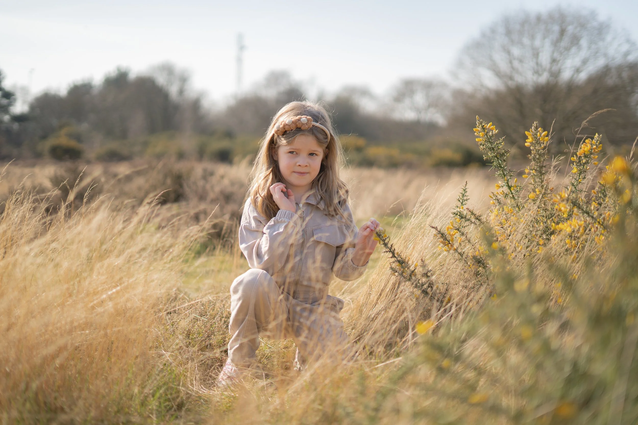 A young girl with long blonde hair, wearing a beige jacket and matching pants, crouching in a field of tall dry grass and yellow flowering bushes, with trees and a cloudy sky in the background.