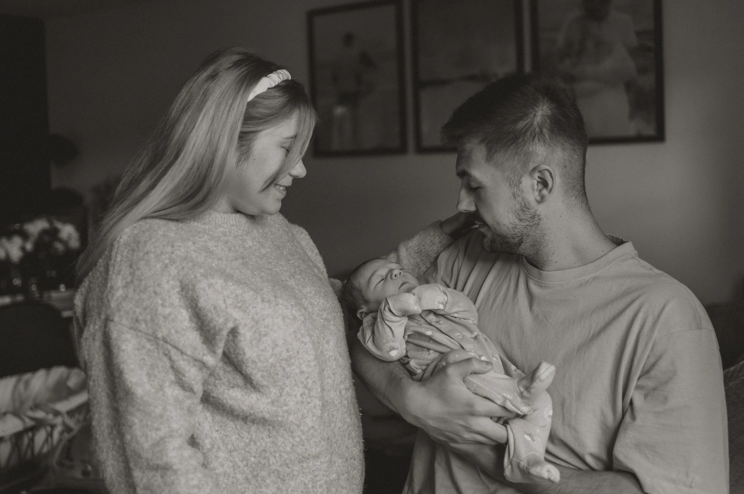 A family of three, a woman, a man, and a newborn, in a warmly lit room. The woman smiles and looks at the man, who is holding the newborn in his arms. The woman gently touches the newborn's forehead. The scene exudes happiness and tenderness.