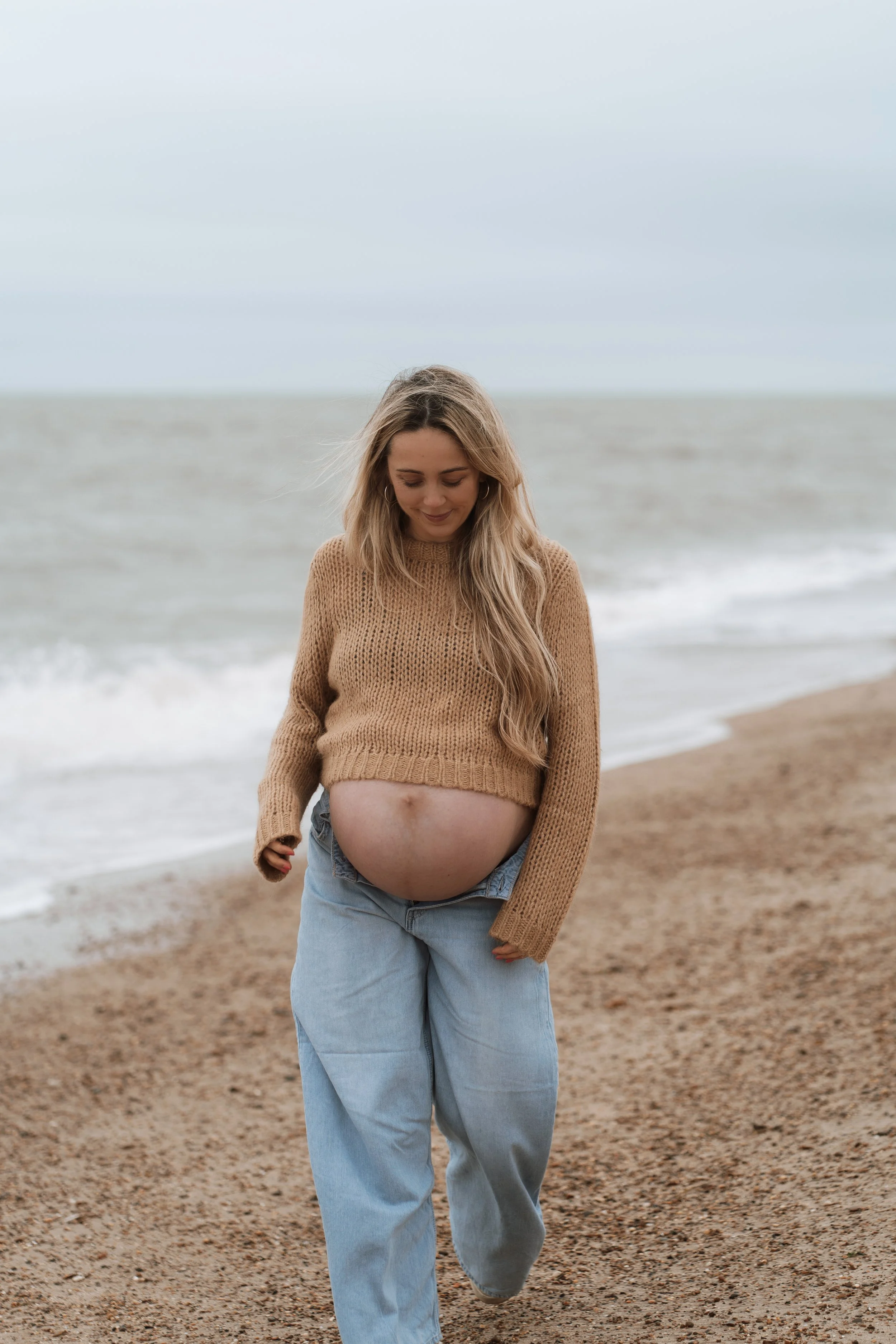 Pregnant woman walking on the beach, smiling, wearing a beige sweater and blue jeans.