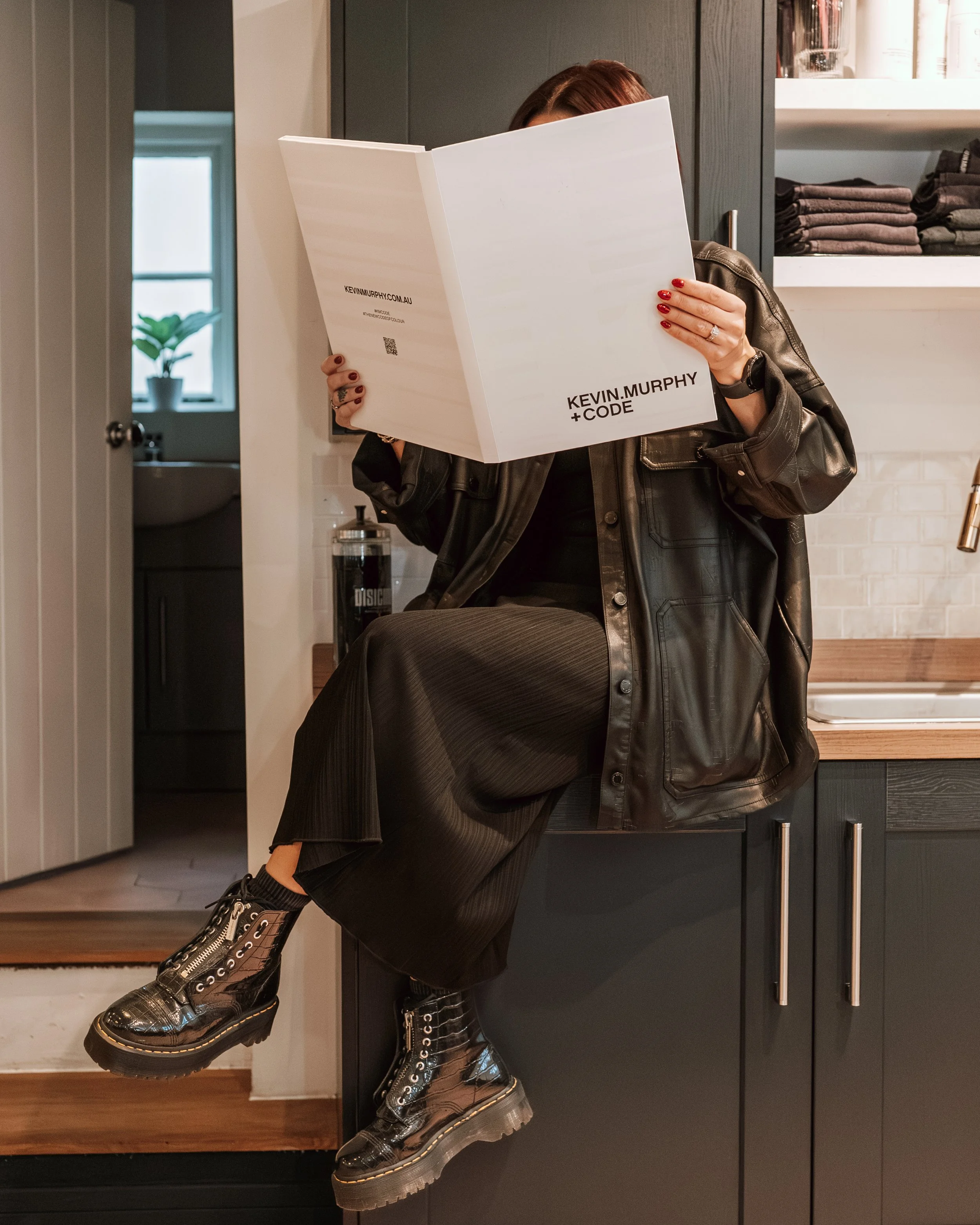 A woman with red hair reading a white book with black text and a website URL, wearing a black leather jacket, black pants, black platform boots, sitting on a kitchen counter.