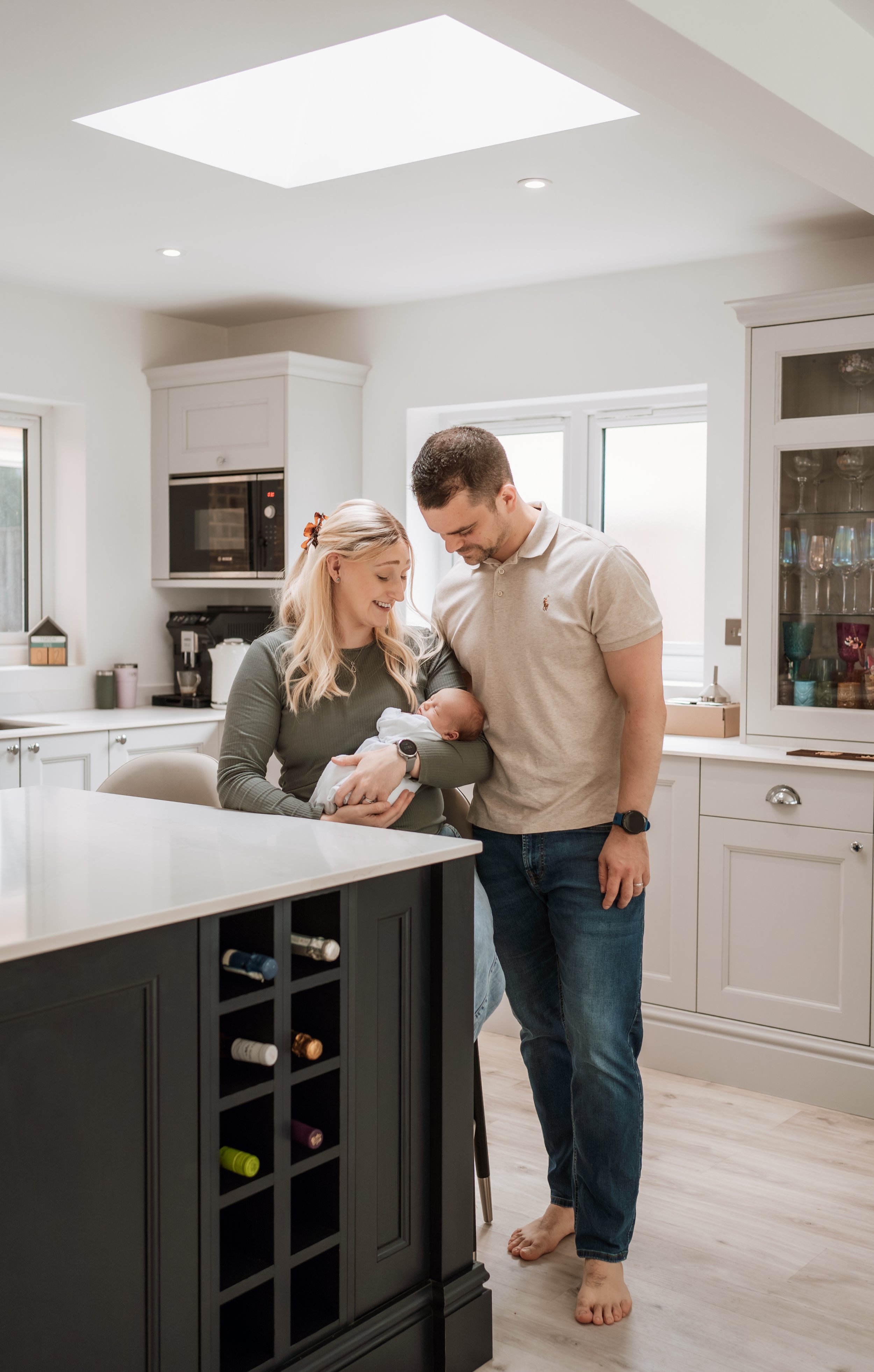 A smiling woman holding a sleeping baby in a kitchen with a man leaning in to look at the baby. The kitchen has white cabinets, a window, and a black wine rack.