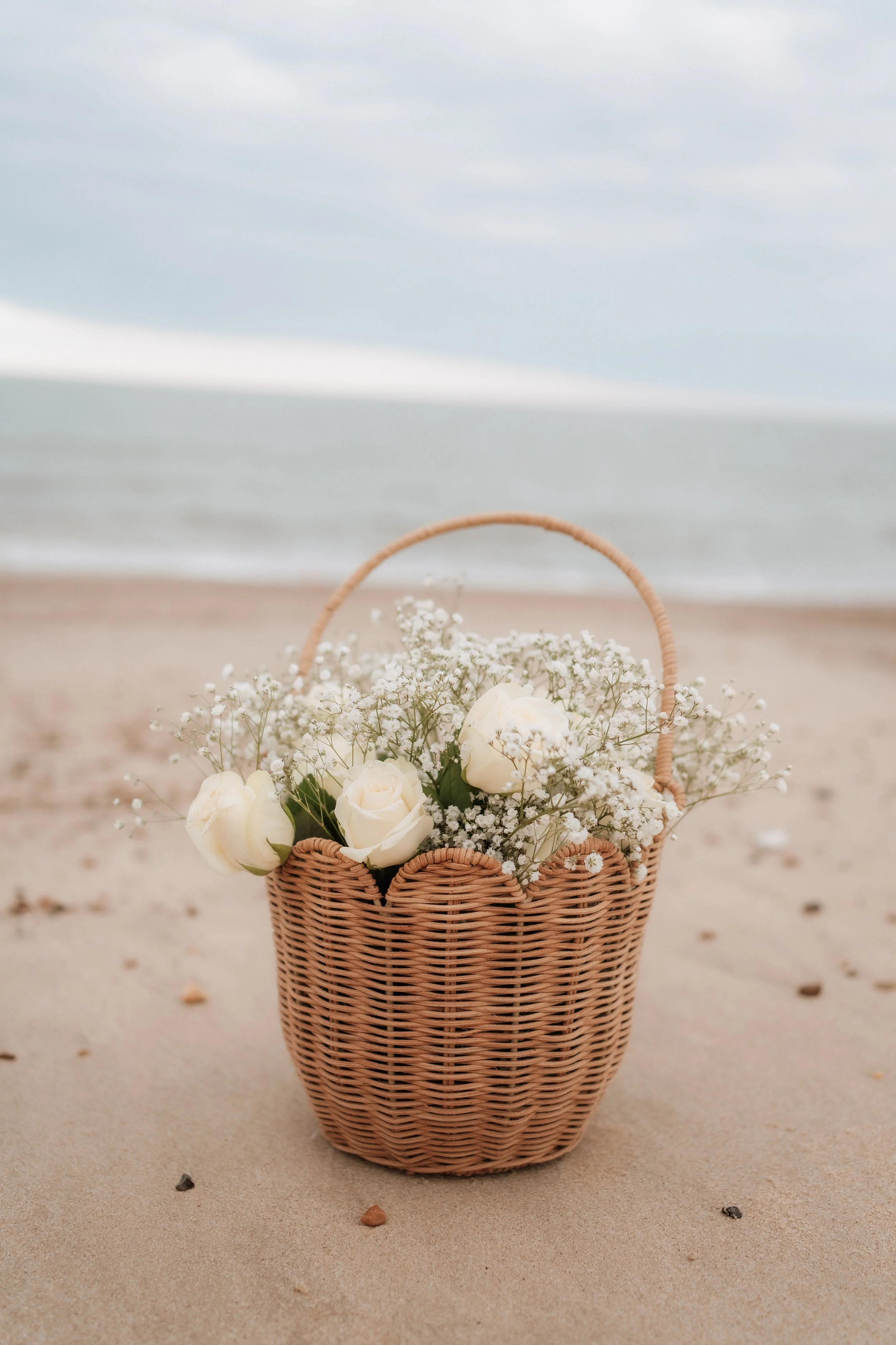A wicker basket filled with white roses and baby's breath flowers on a sandy beach with the ocean and cloudy sky in the background.