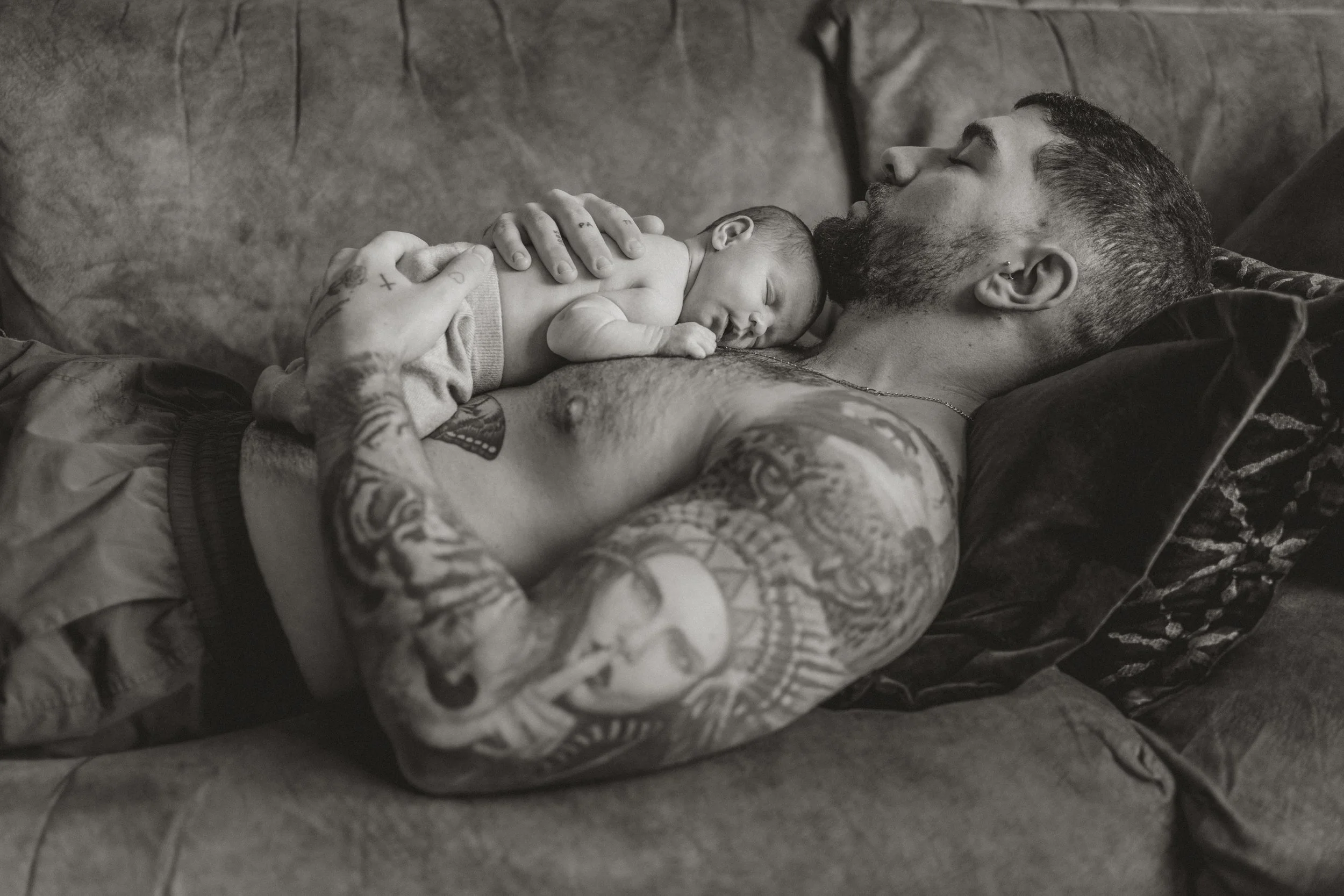 A bearded man with tattoos on his arm and chest lying on a couch, holding a sleeping baby on his chest, with eyes closed.