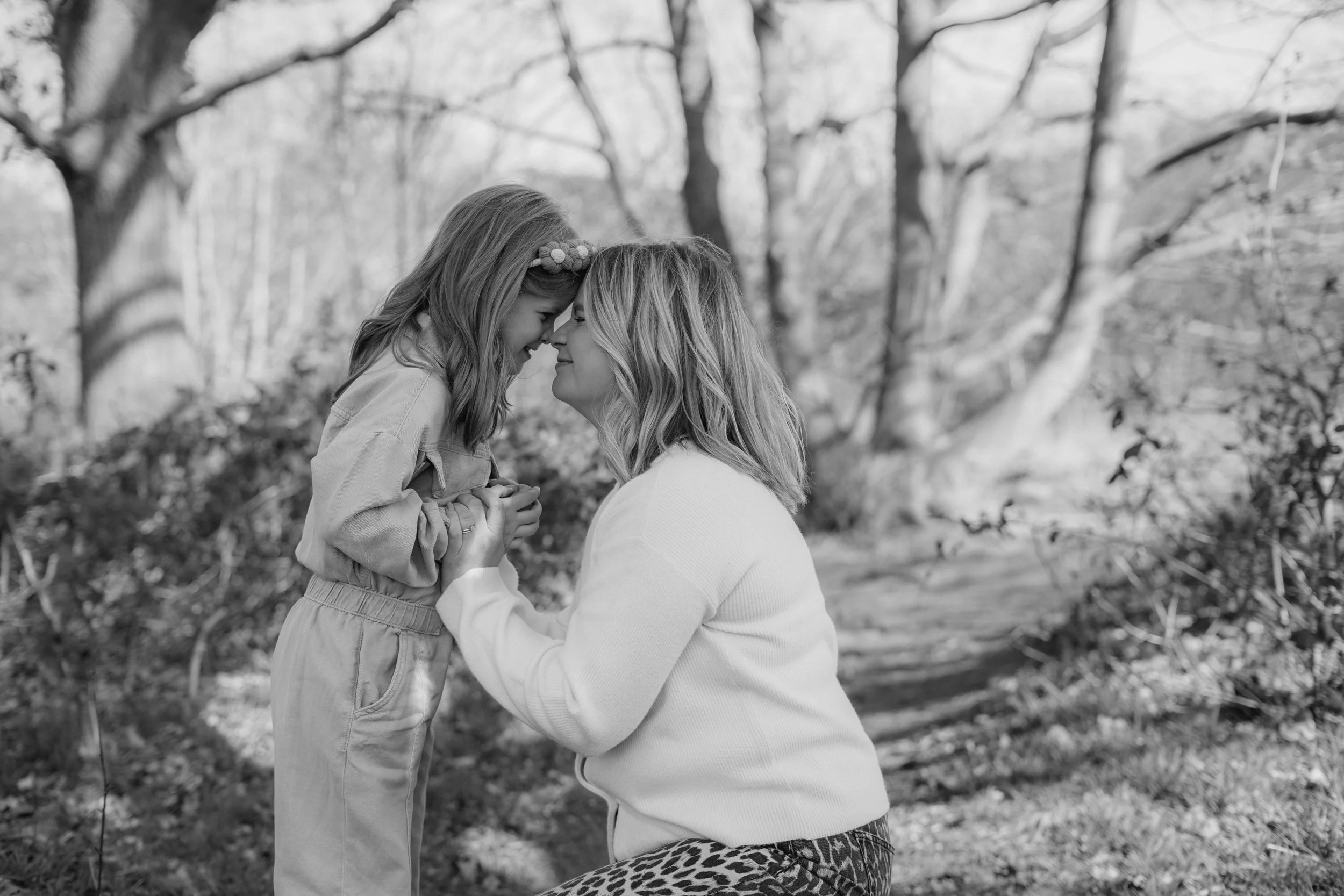 A woman and a young girl are touching foreheads and smiling at each other outdoors in a wooded area.