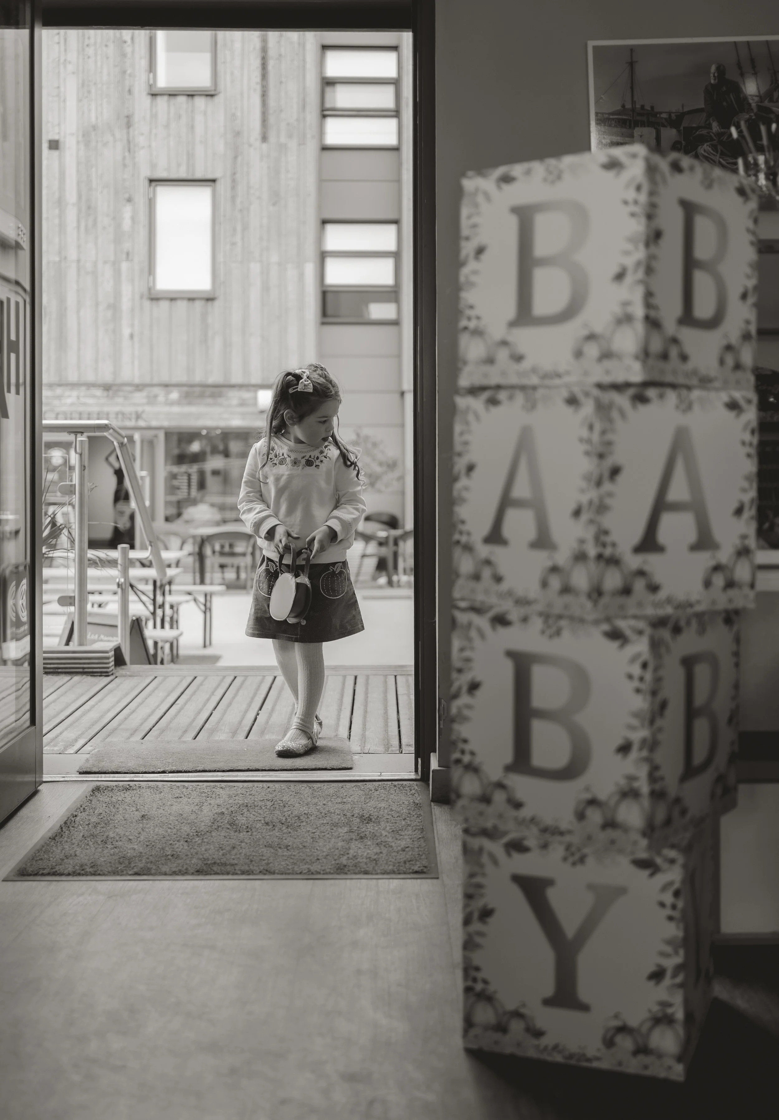 A young girl standing outside a cafe or shop door, holding a small bag, with a modern building in the background. Inside, stacked blocks with letters B, A, B, Y are visible, indicating it might be a celebration or themed decor.