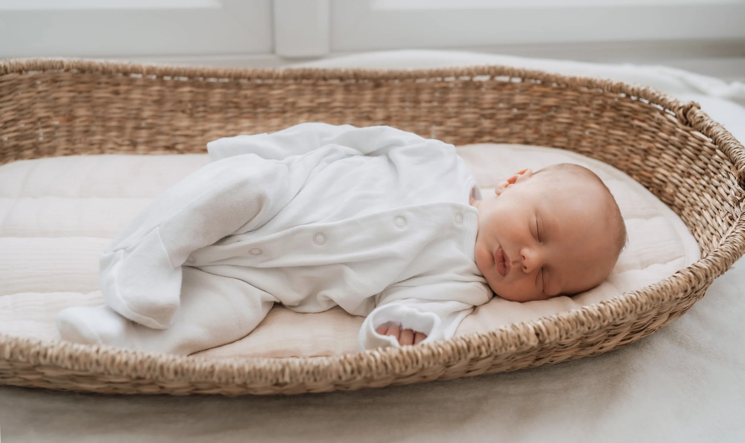 A sleeping baby in a white onesie lying on a padded mattress in a wicker bassinet.