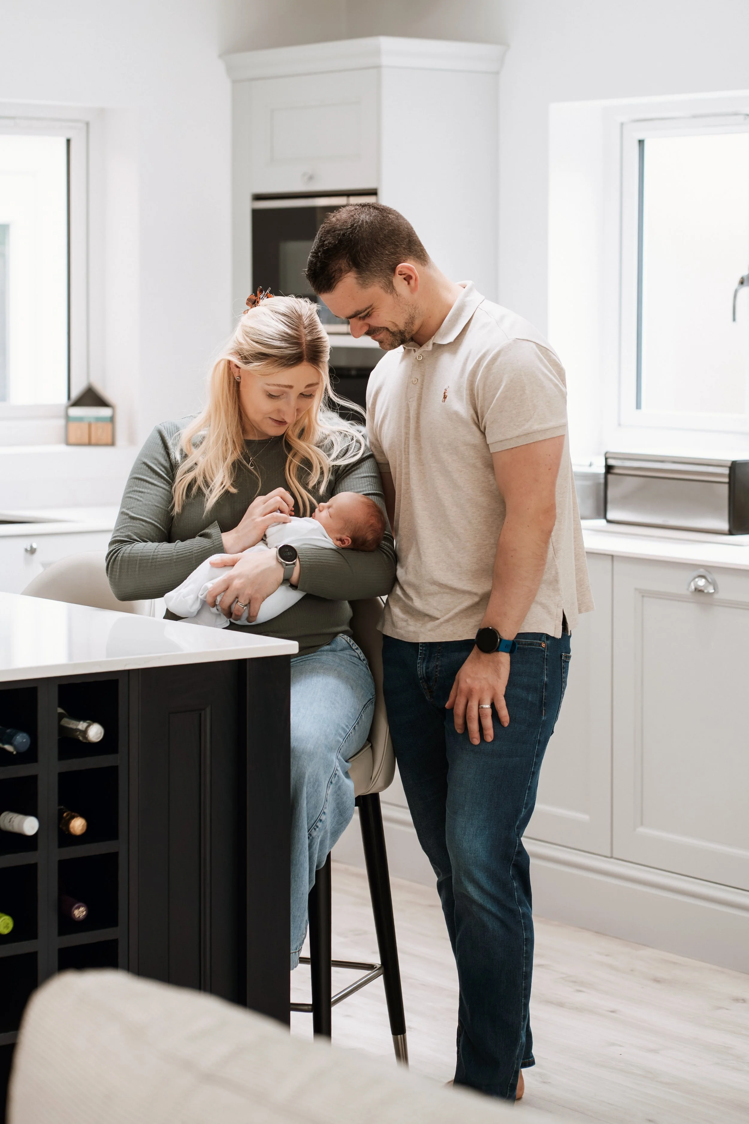 A woman sitting at a kitchen counter holding a newborn baby, with a man standing beside her looking at the baby in her arms.