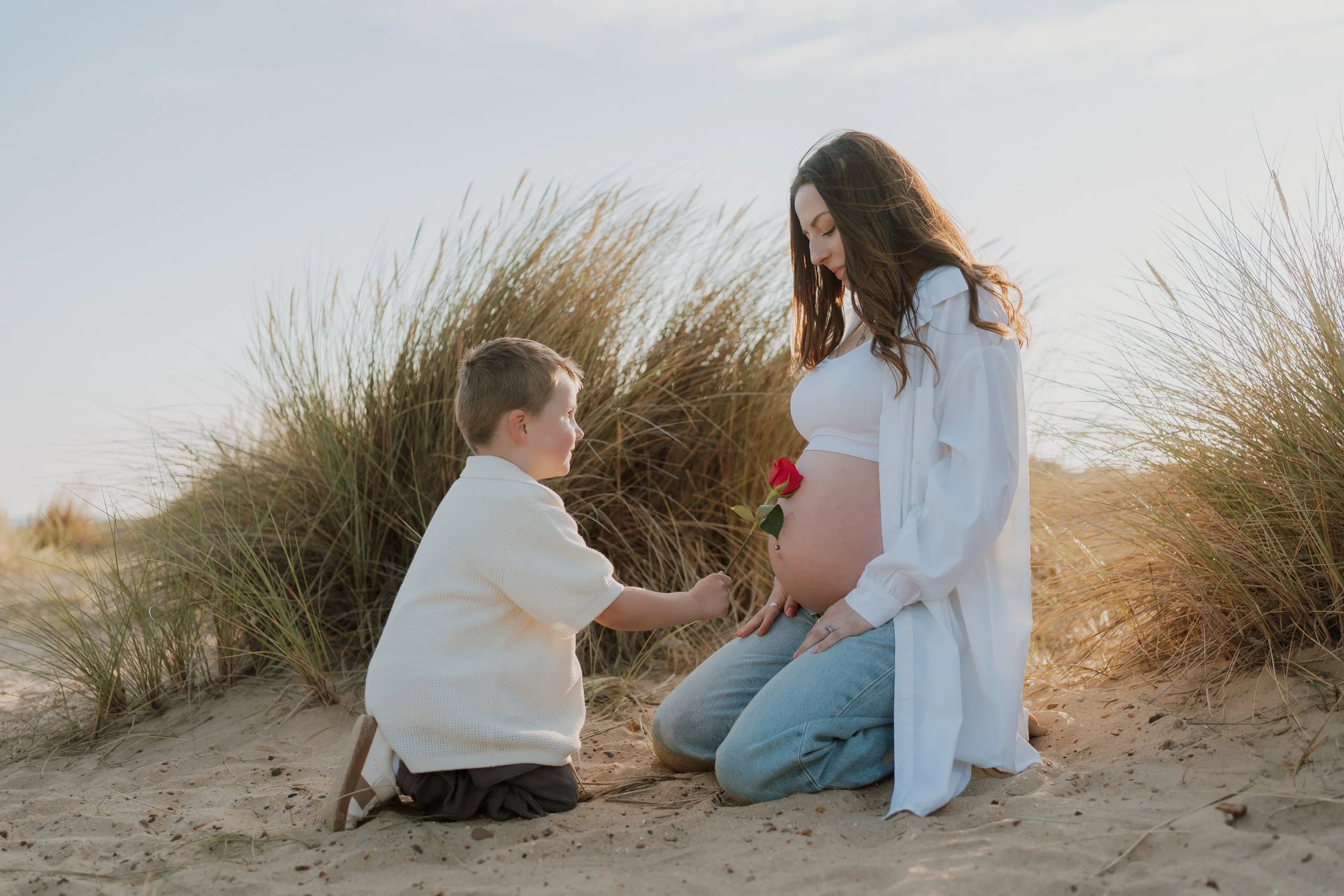 A pregnant woman kneeling on the sand with a red rose on her belly, and a young boy kneeling in front of her, holding the rose and looking at her. They are outdoors on a sandy area with tall grass and a clear sky.