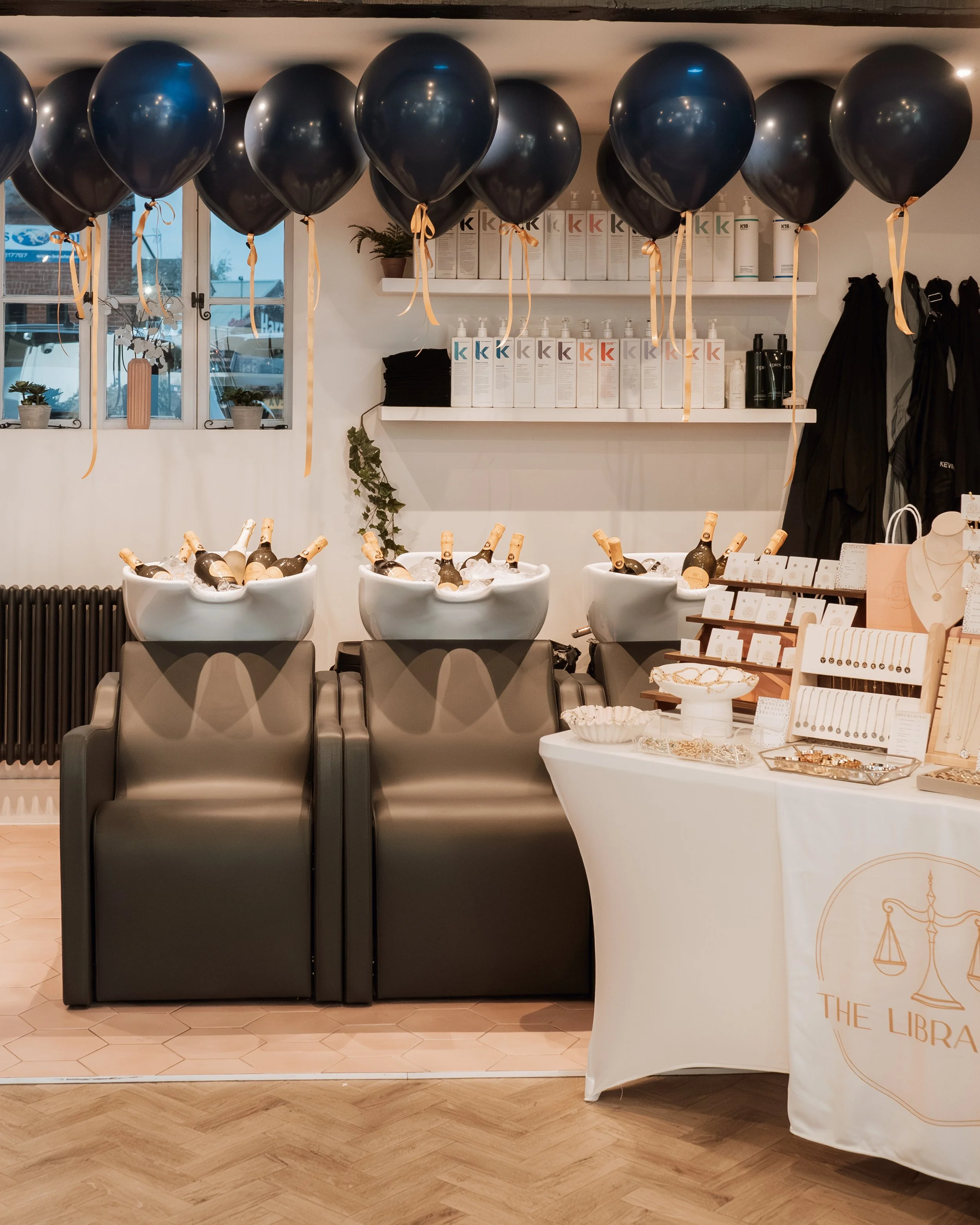 A display with black balloons hanging from the ceiling, bottles of champagne in white buckets, jewelry on a table, and shelves with hair or skincare products.