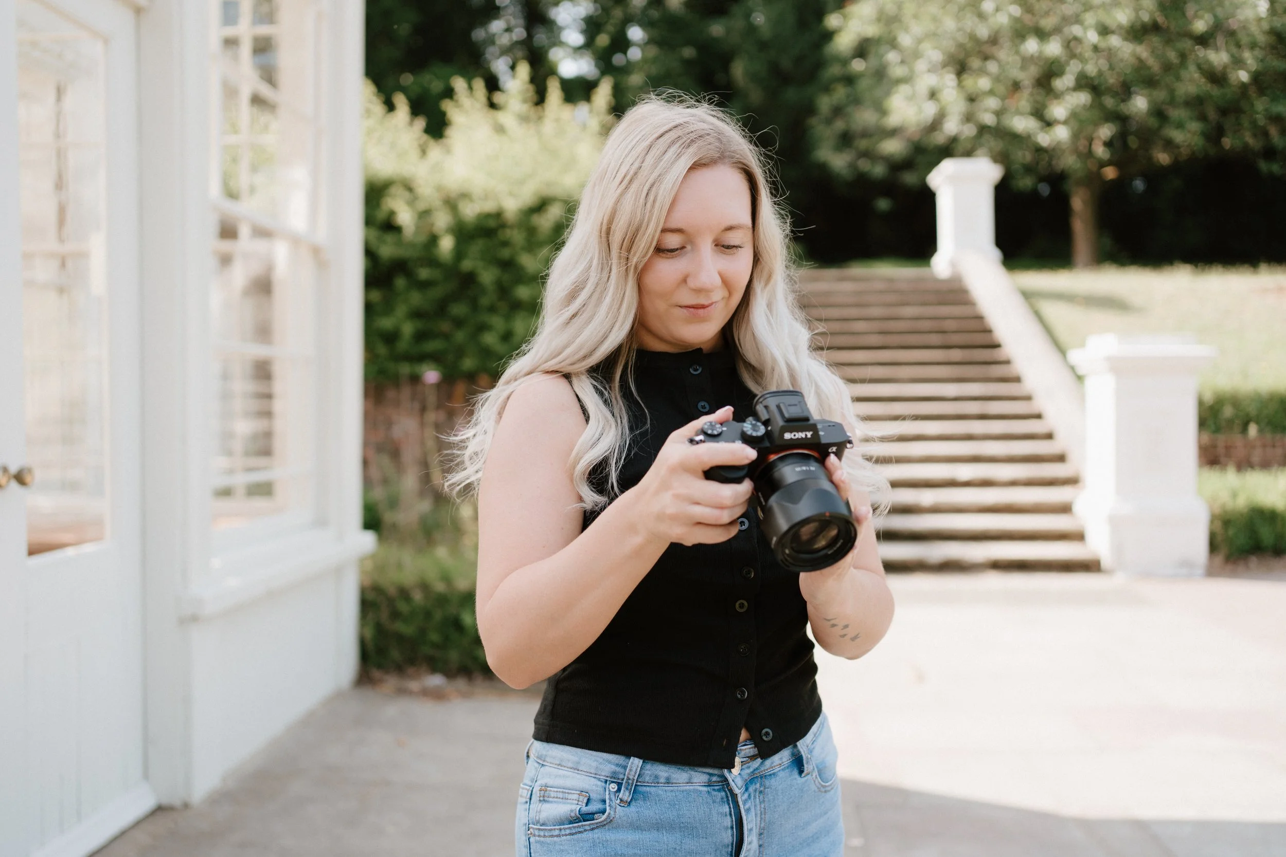 A woman with long blonde hair wearing a black sleeveless top and light blue jeans holding a Sony camera outdoors near a staircase.