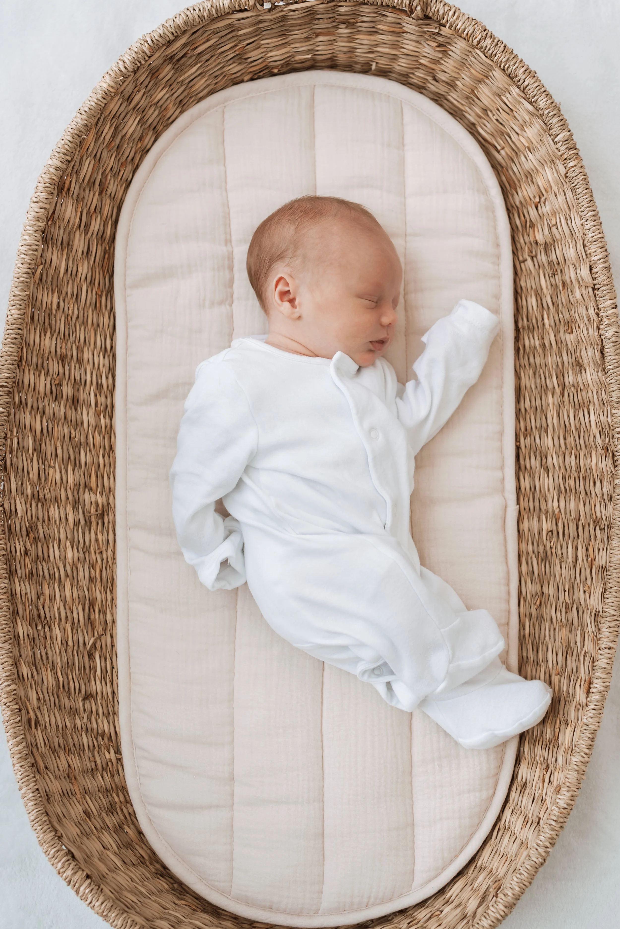 A sleeping baby in a white onesie lying on a beige and white cushioned mattress inside a woven wicker bassinet.