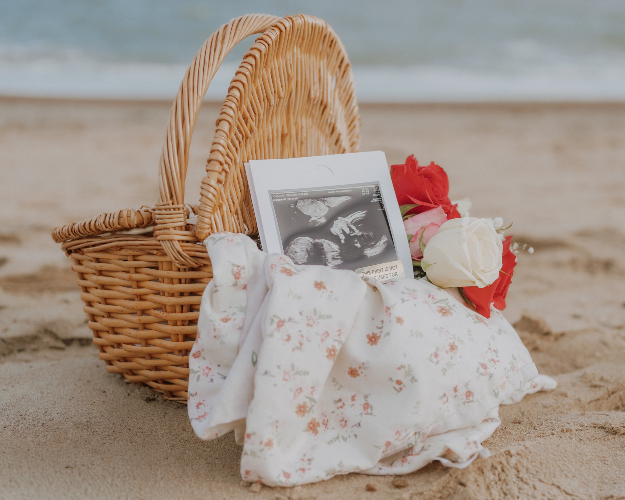 A wicker basket on the sand at the beach with an ultrasound photo, a pillow with a floral pattern, and a bouquet of red, pink, and white roses.