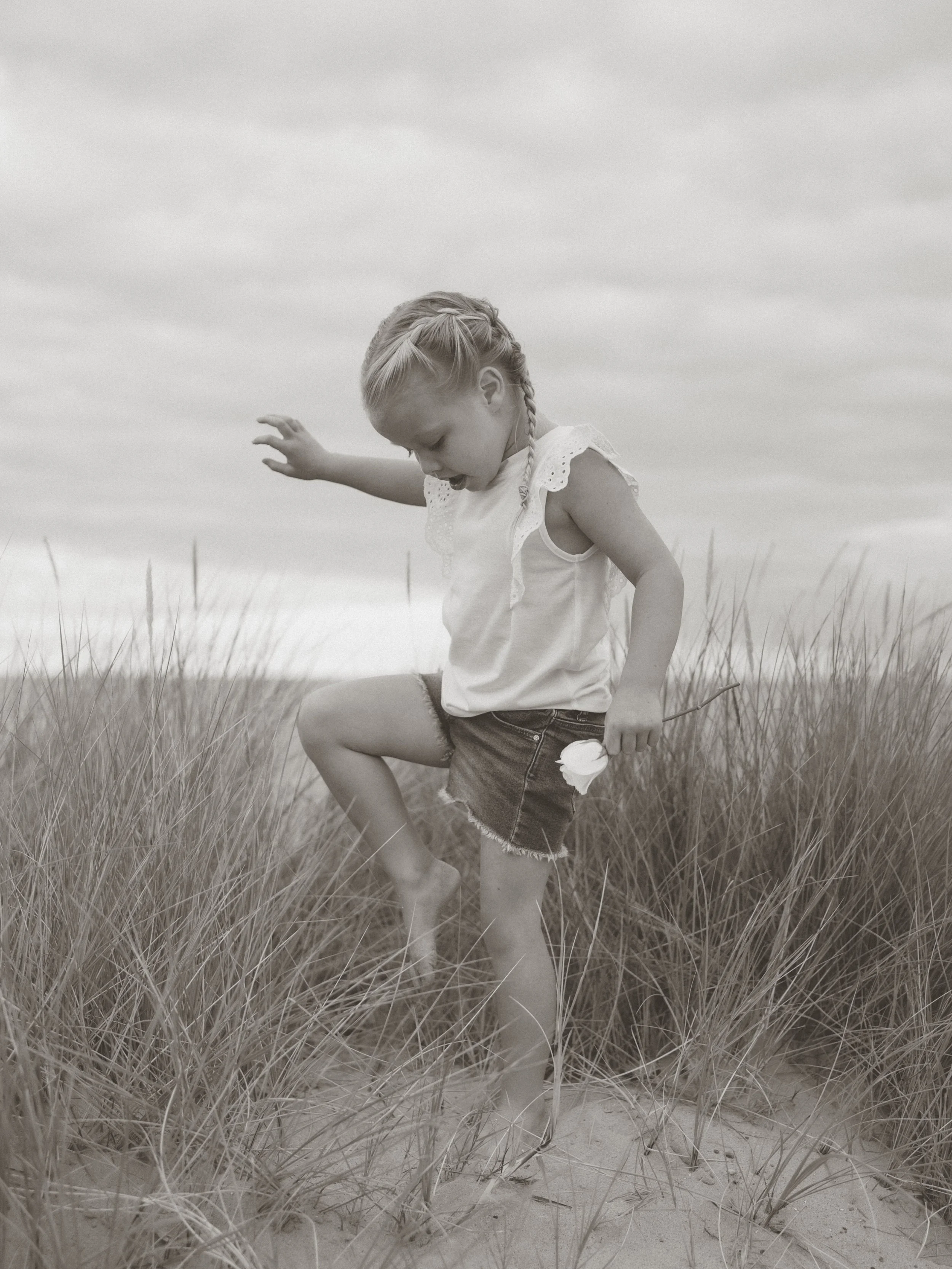 A young girl with braided hair wearing a sleeveless top and denim shorts balances on one foot in a grassy sand dune, with a cloudy sky in the background.